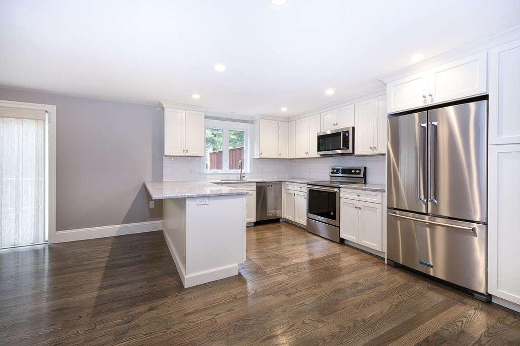 Modern white kitchen with stainless-steel appliances, a stone-topped peninsula, and dark wood floors.