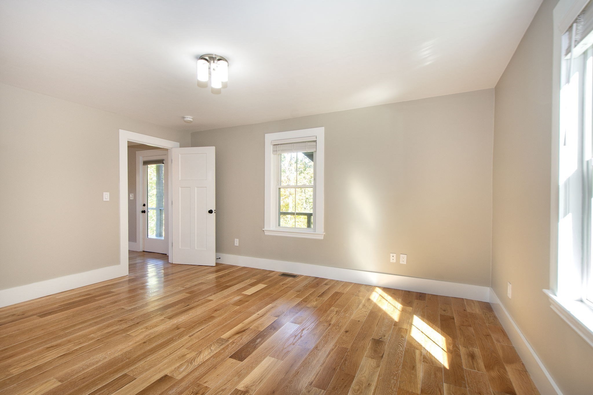 Empty room with hardwood floors, beige walls, and sunlight streaming through the windows.