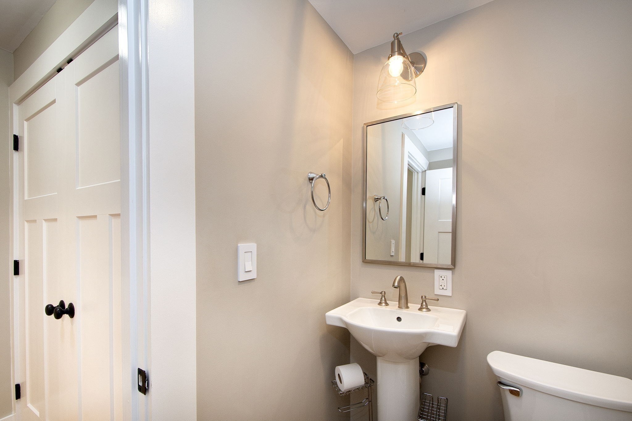Bathroom with a pedestal sink, rectangular mirror, and partially visible toilet beside a paneled door.