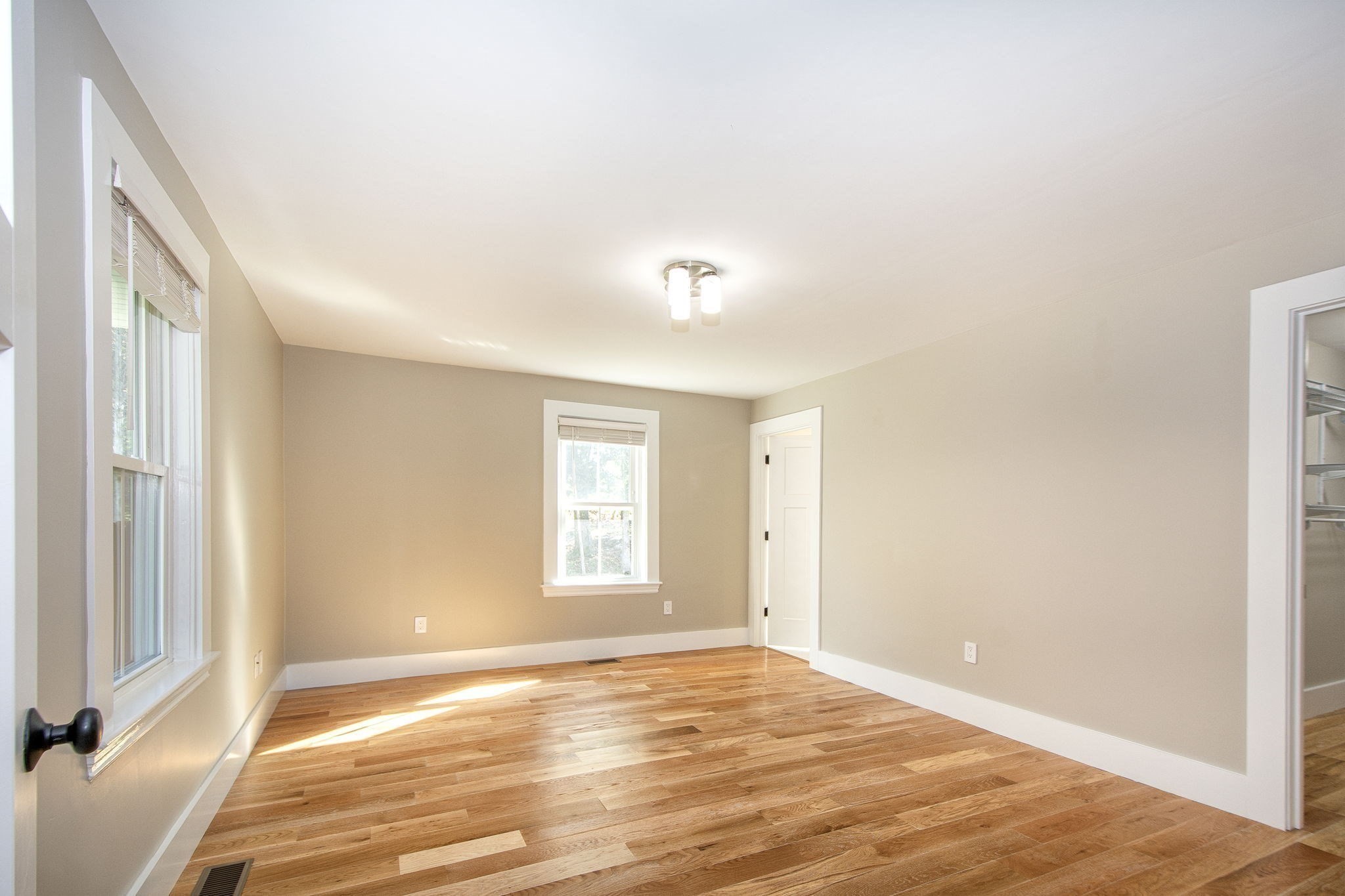 Empty bedroom with hardwood floors, beige walls, and windows letting in sunlight.