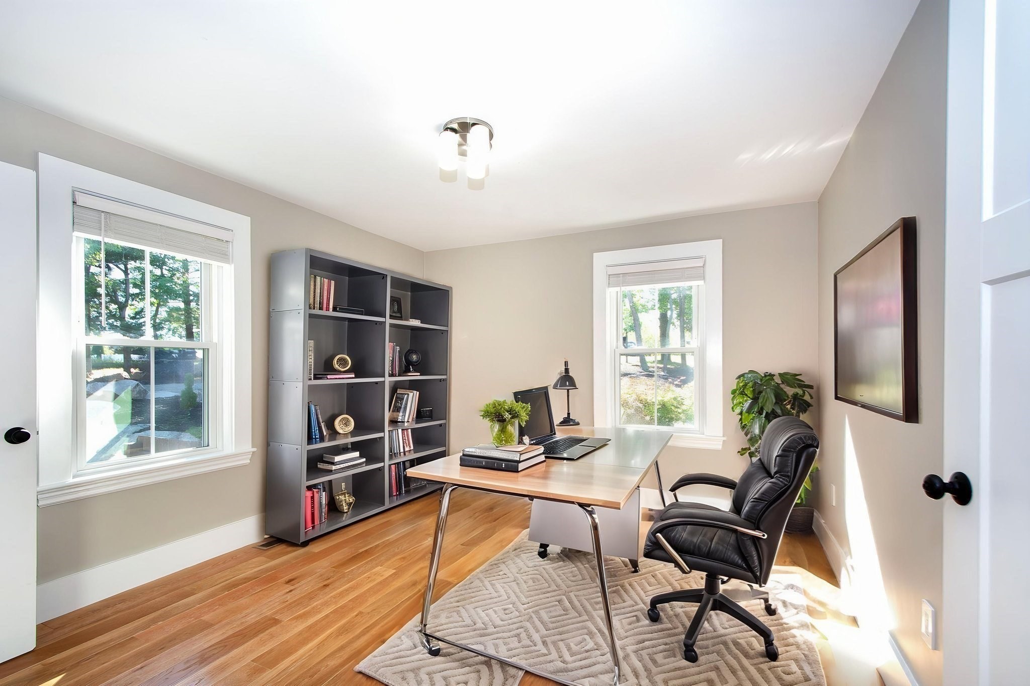 Bright home office with a desk and laptop, black bookcase, leather chair, and hardwood floors.