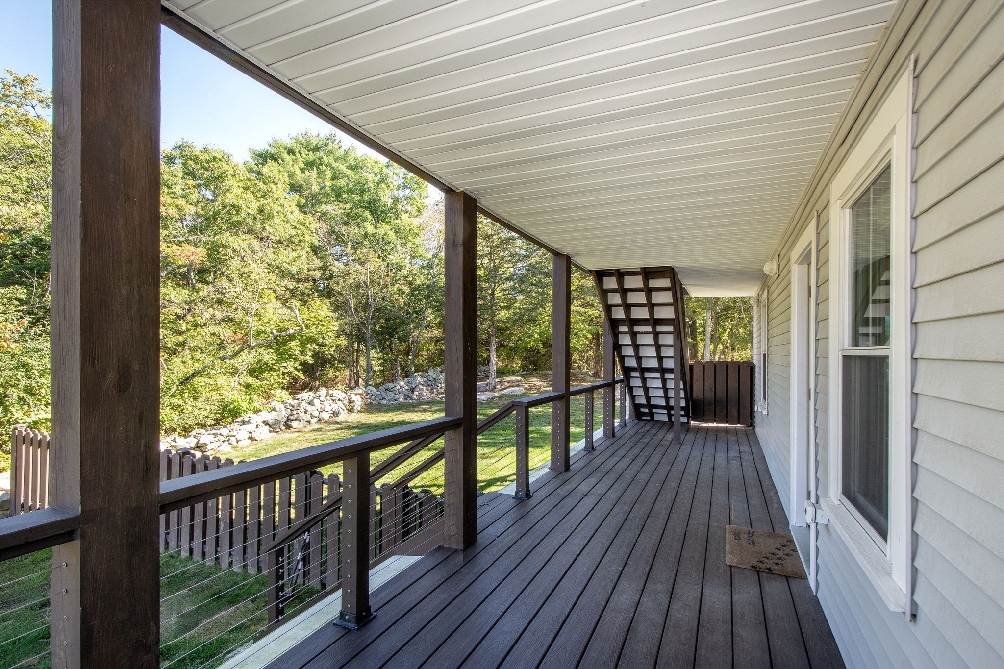 Covered deck with dark wood flooring and cable railings overlooking a wooded yard and stone wall.