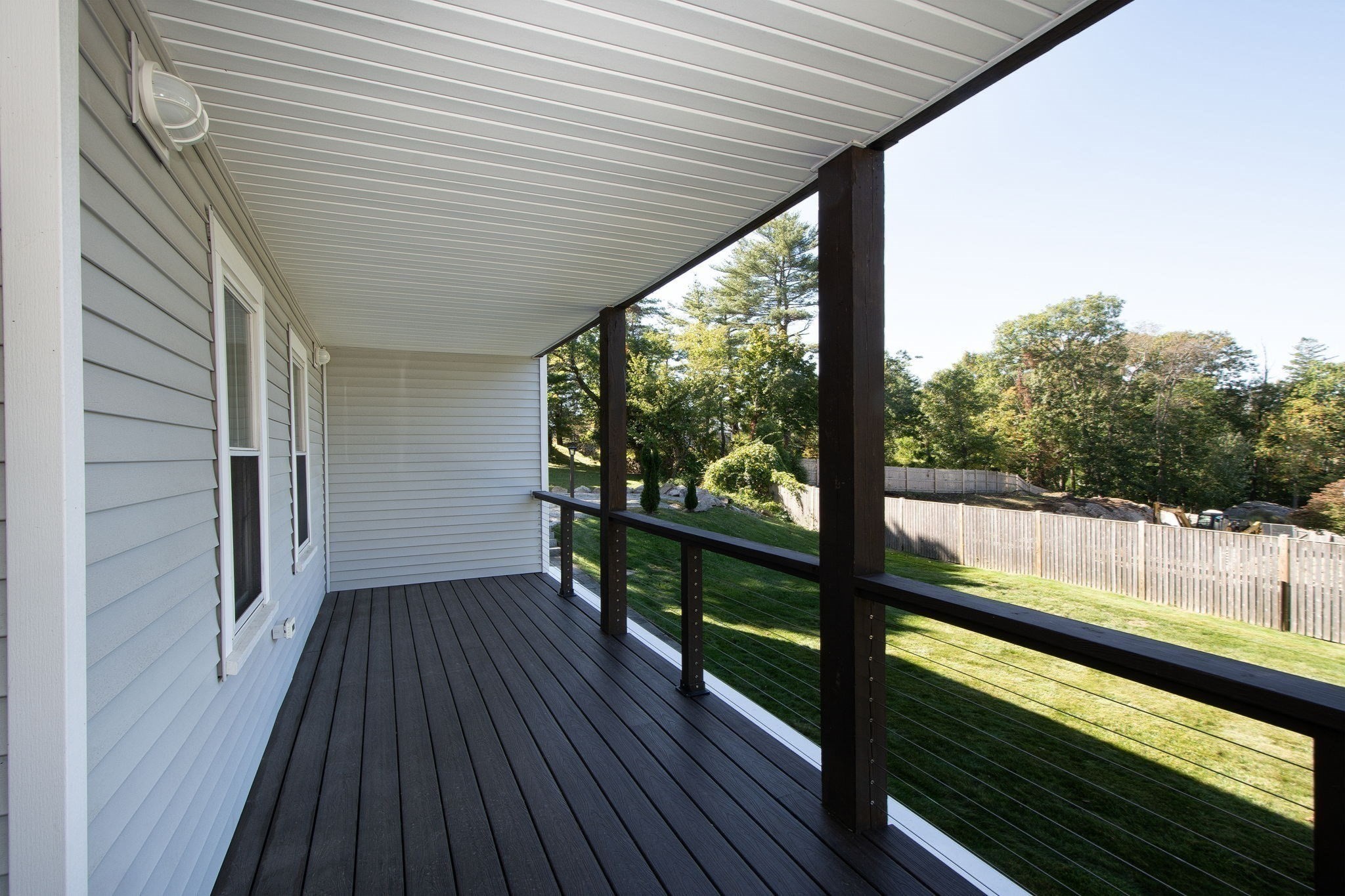 Covered deck with dark flooring and cable railing overlooking a fenced, tree-lined lawn.