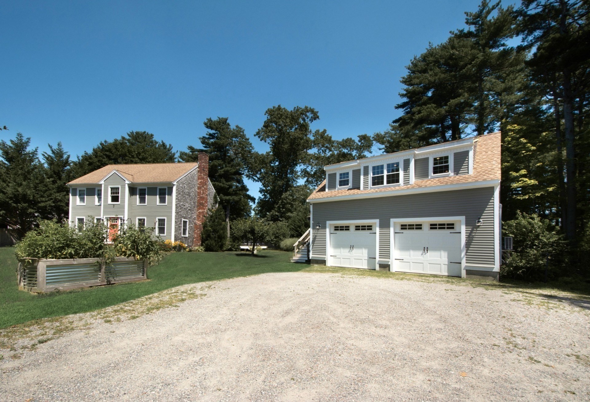 Gray two-story home and detached two-door garage beside a gravel driveway and raised garden bed.