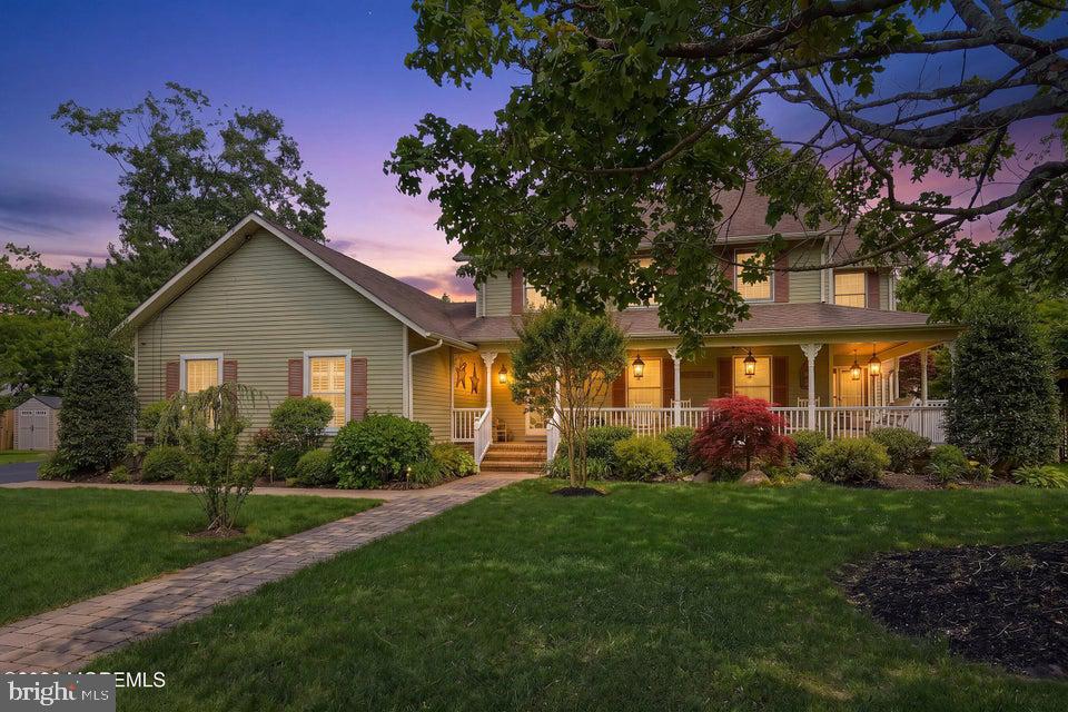 Green-sided house with an illuminated wraparound porch and brick walkway at dusk.