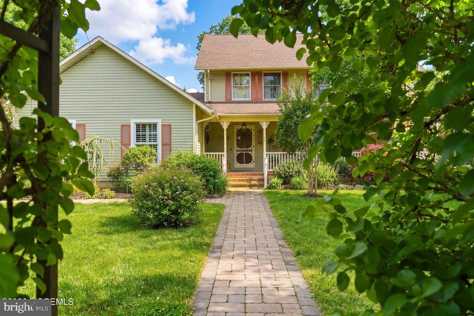 Two-story home with a covered porch and paved walkway through a landscaped lawn, framed by leafy branches.