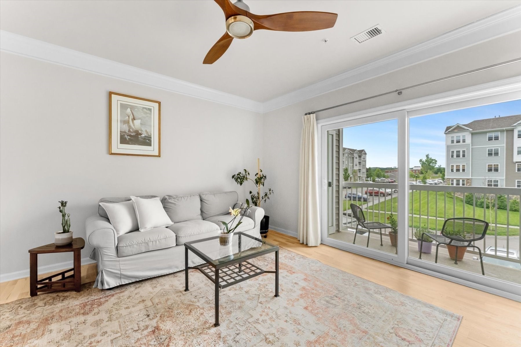 Furnished living room with a white sofa and sliding doors opening onto a balcony overlooking apartment grounds.