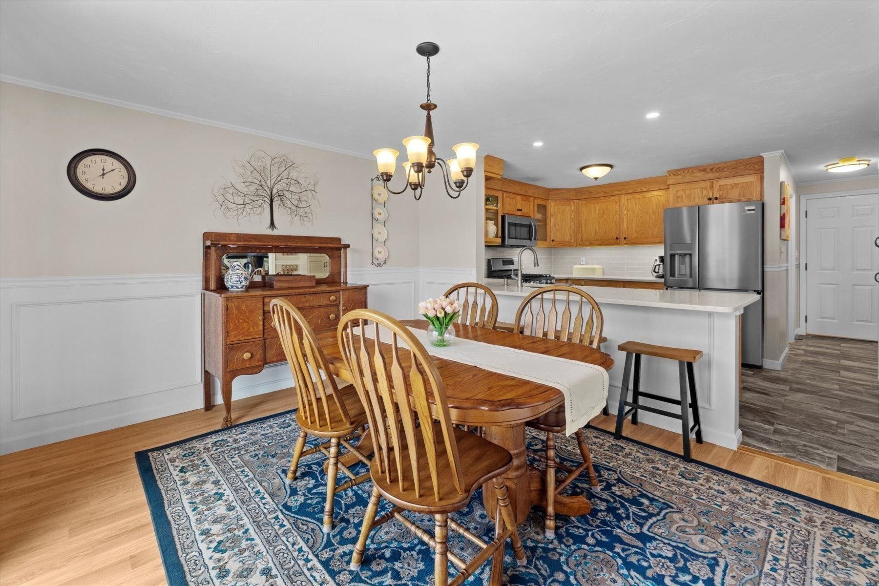 Dining area with a wooden table on a blue patterned rug beside an oak-cabinet kitchen.