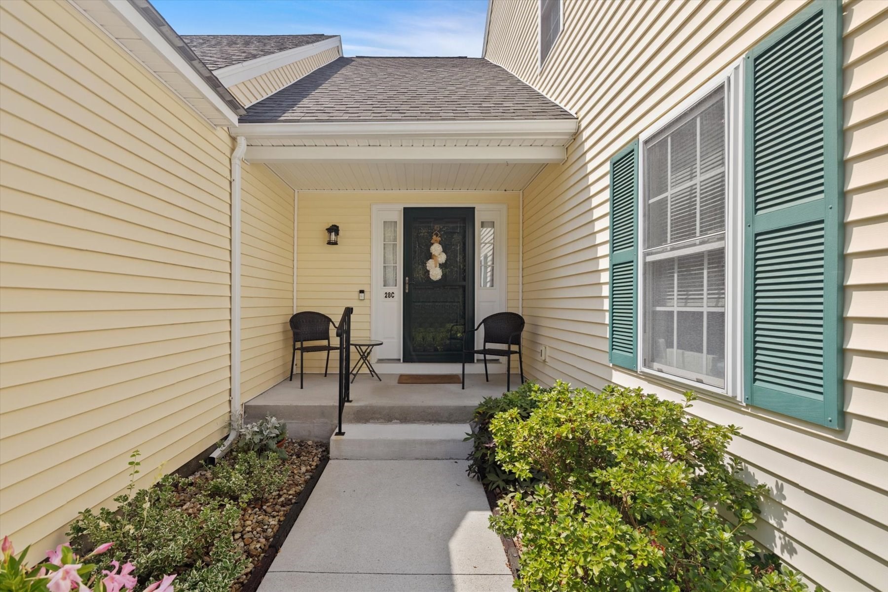 Covered home entrance with a concrete walkway, green shutters, and chairs beside the door.