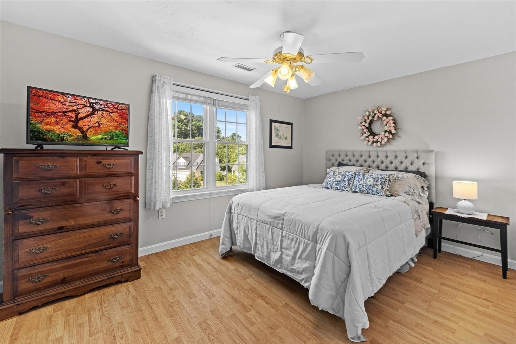 Bedroom with gray tufted headboard, white bedding, large curtained window, and dark wood dresser.