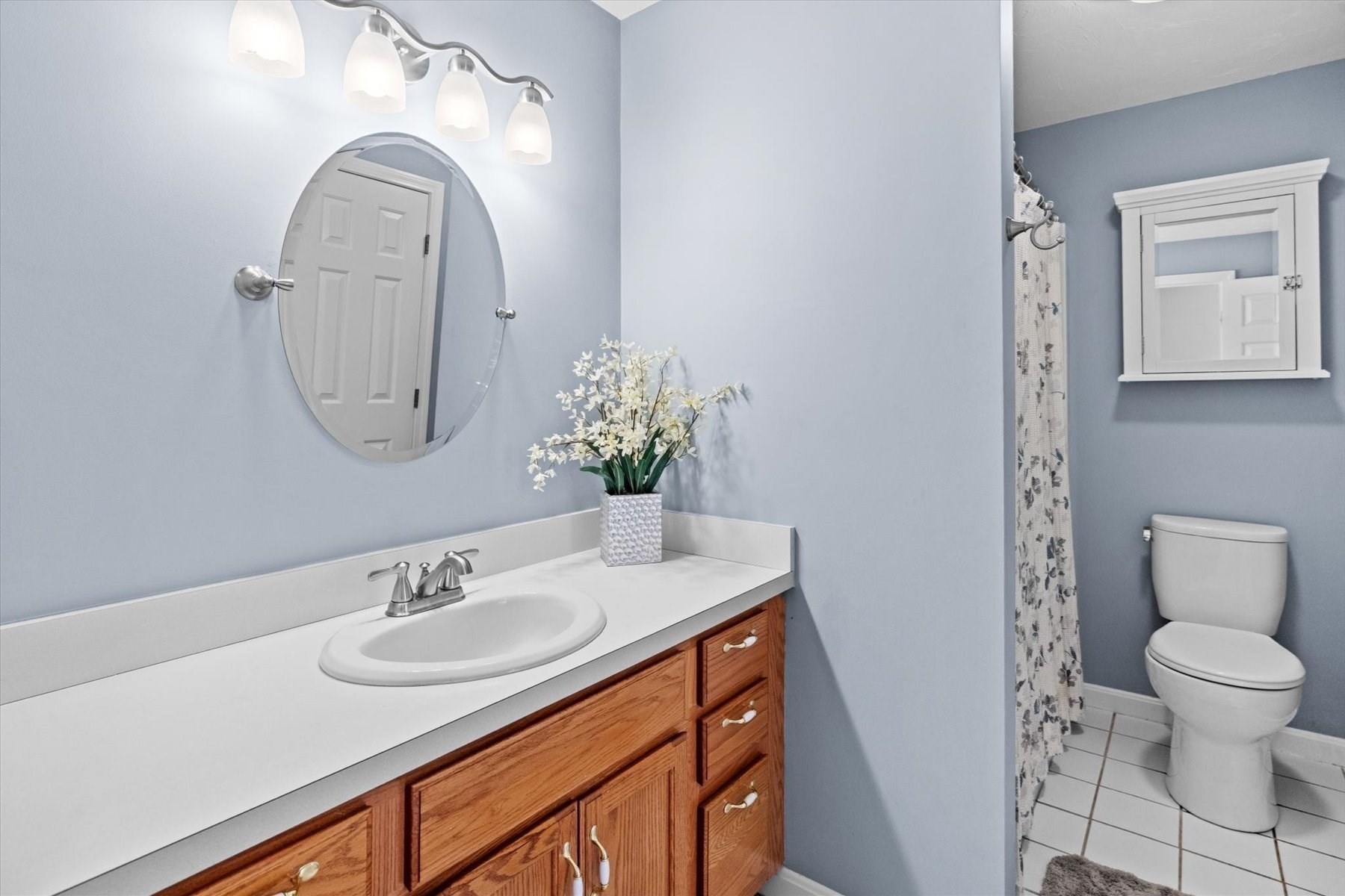 Bathroom with a wood vanity and oval mirror beside a toilet and patterned shower curtain.