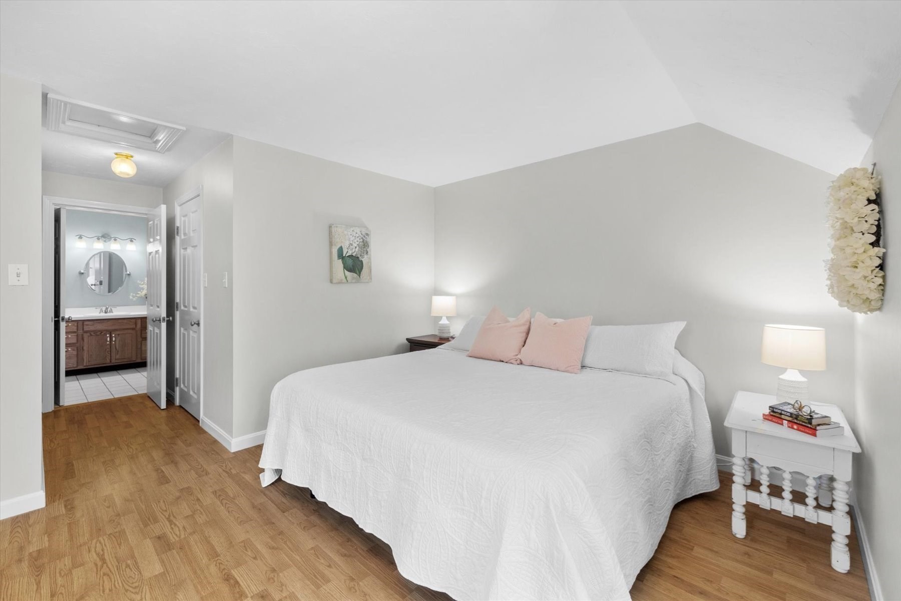 Bedroom with a white bed, pink accent pillows, wood flooring, and an adjoining bathroom.
