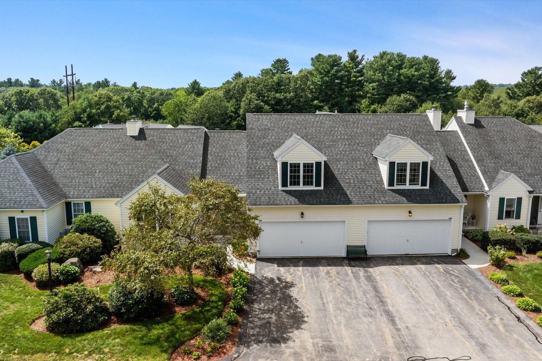 Attached cream-sided home with two garage doors, dormer windows, landscaped grounds, and woods behind it.