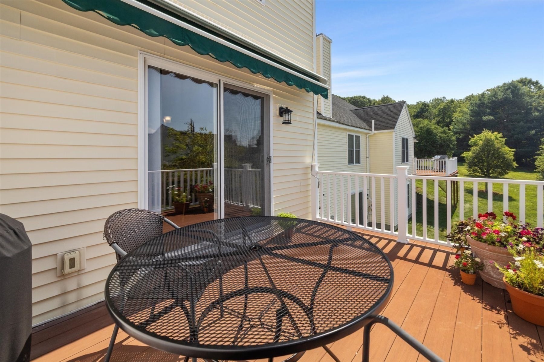 Deck with a round patio table, flower pots, and a green awning overlooking lawn and trees.