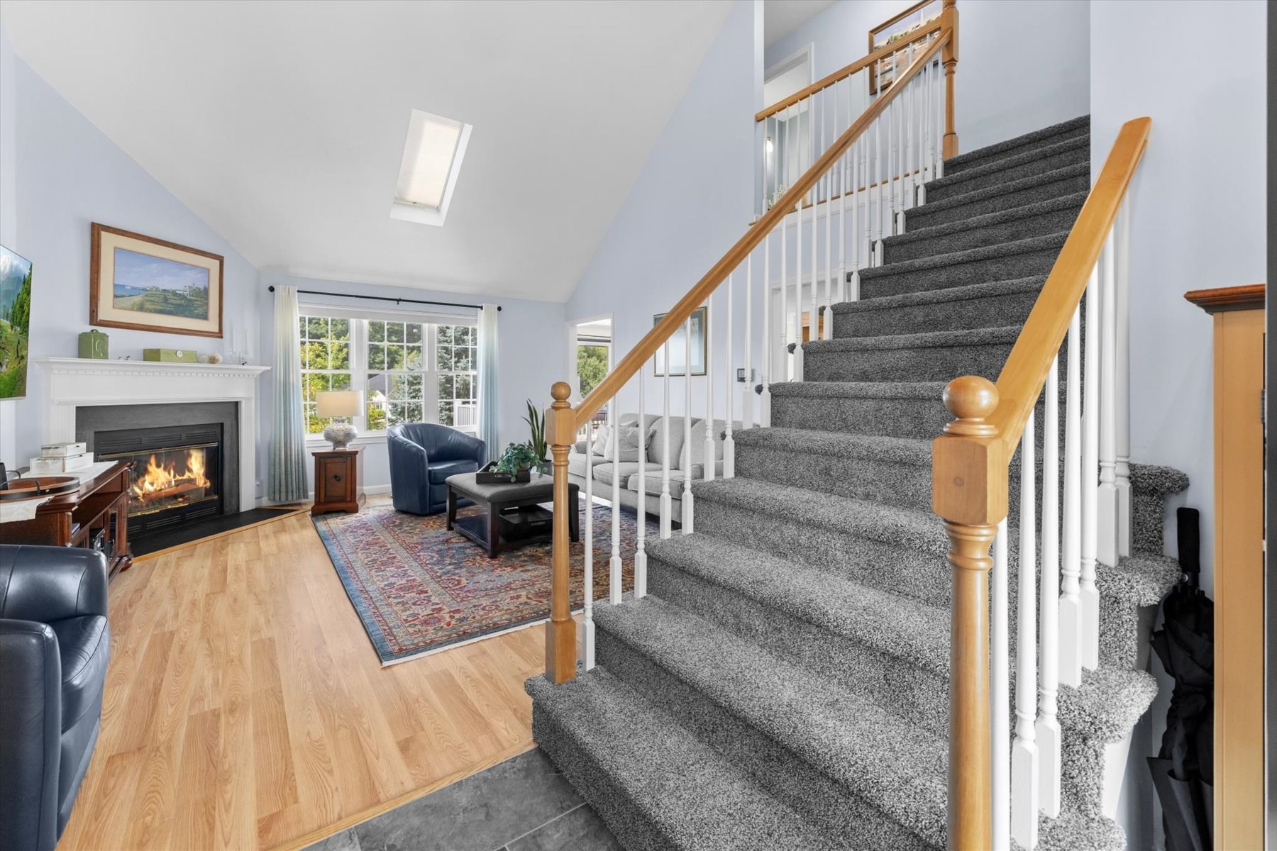 Furnished living room with a fireplace beside a carpeted staircase and bright, large windows.