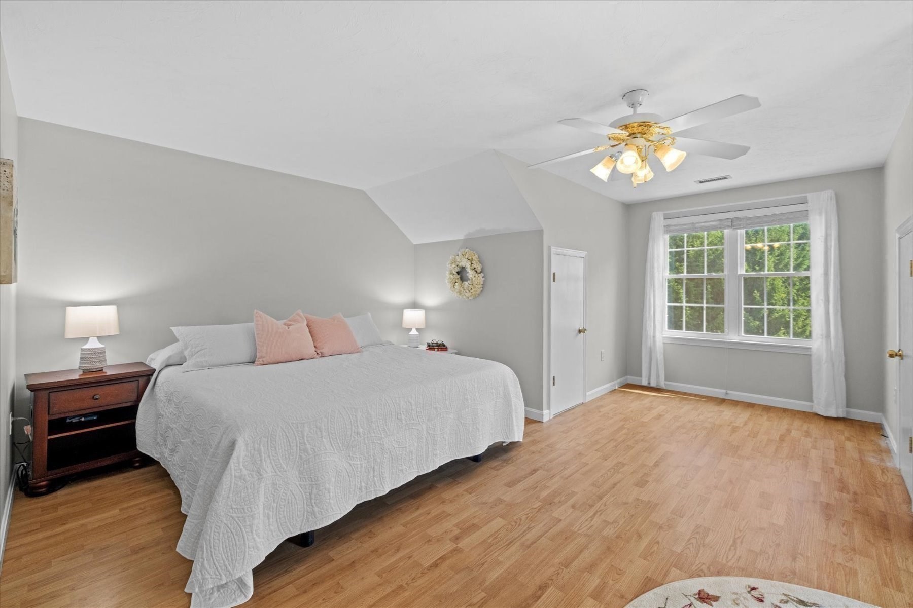 Bedroom with a white-quilted bed, blush pillows, light wood floors, and a large window.