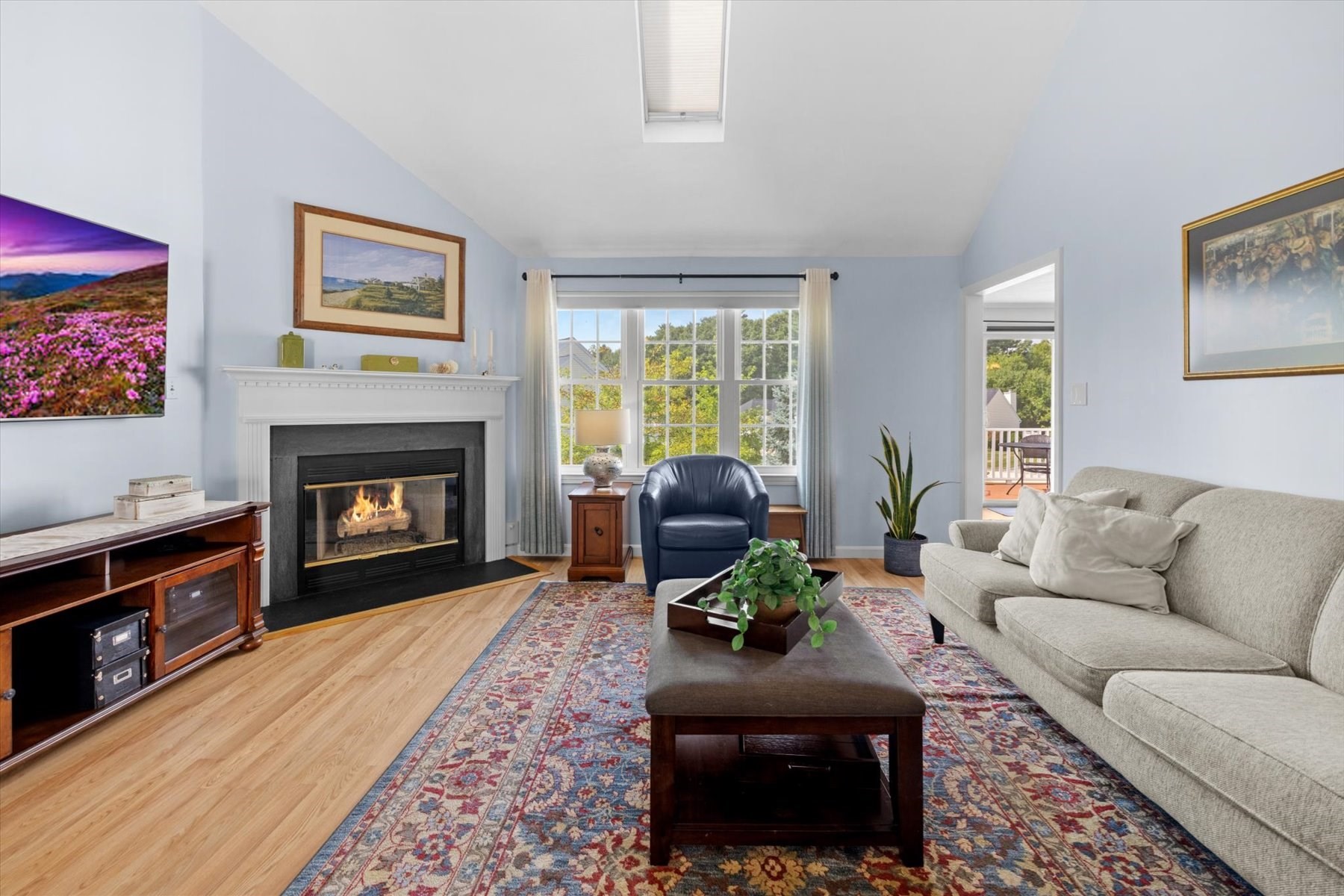 Living room with a fireplace, vaulted ceiling, large windows, and a doorway to a deck.