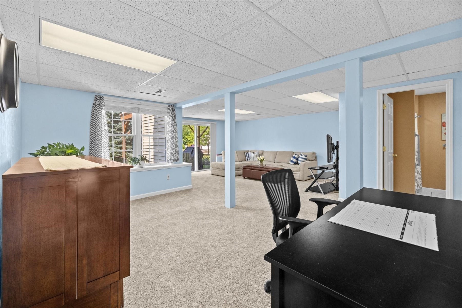 Home office opening onto a sitting area with a beige sectional and large windows.