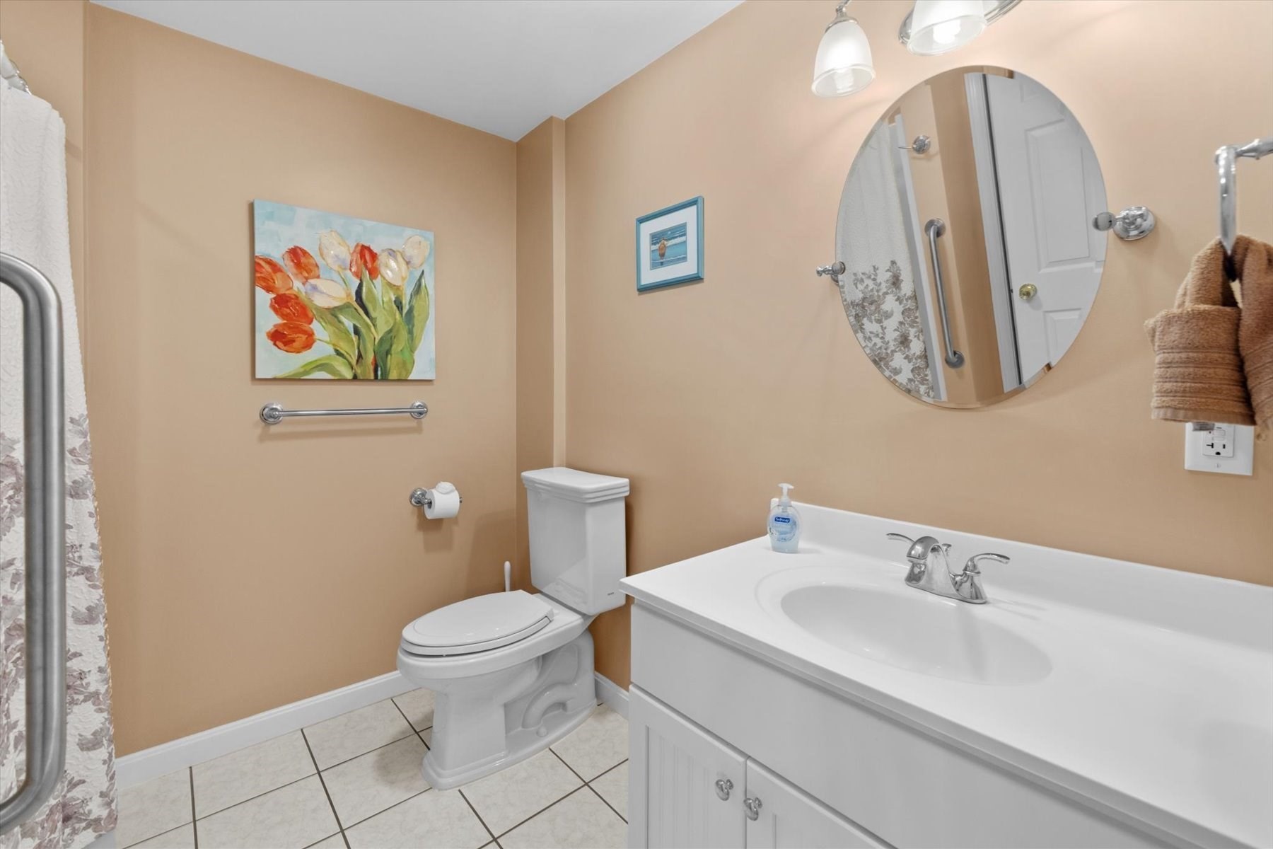 Bathroom with a white vanity and toilet, tiled floor, oval mirror, and shower grab bar.