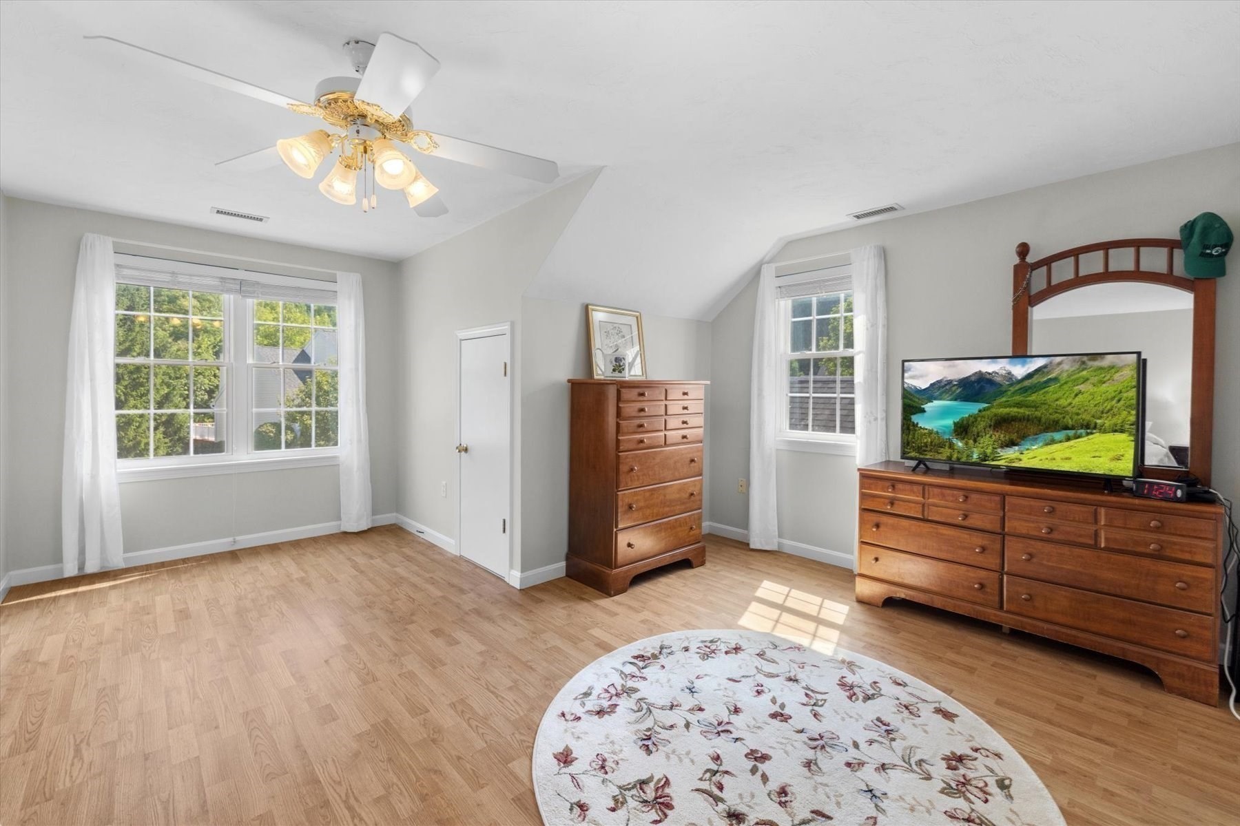 Bright bedroom with sloped ceiling, wooden dressers, TV, and floral rug.