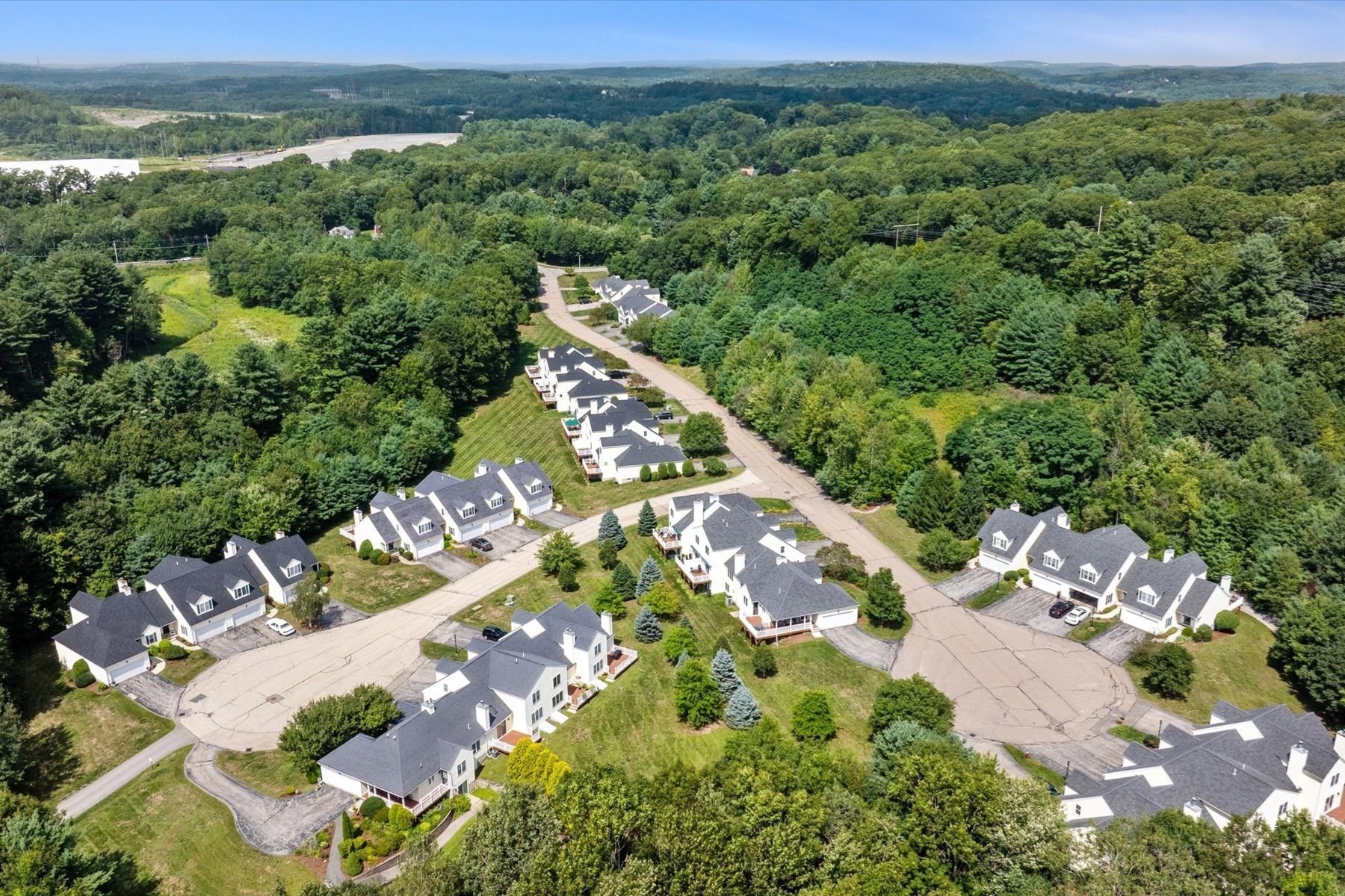 Aerial view of white homes along a curving road amid dense woodland and rolling hills.