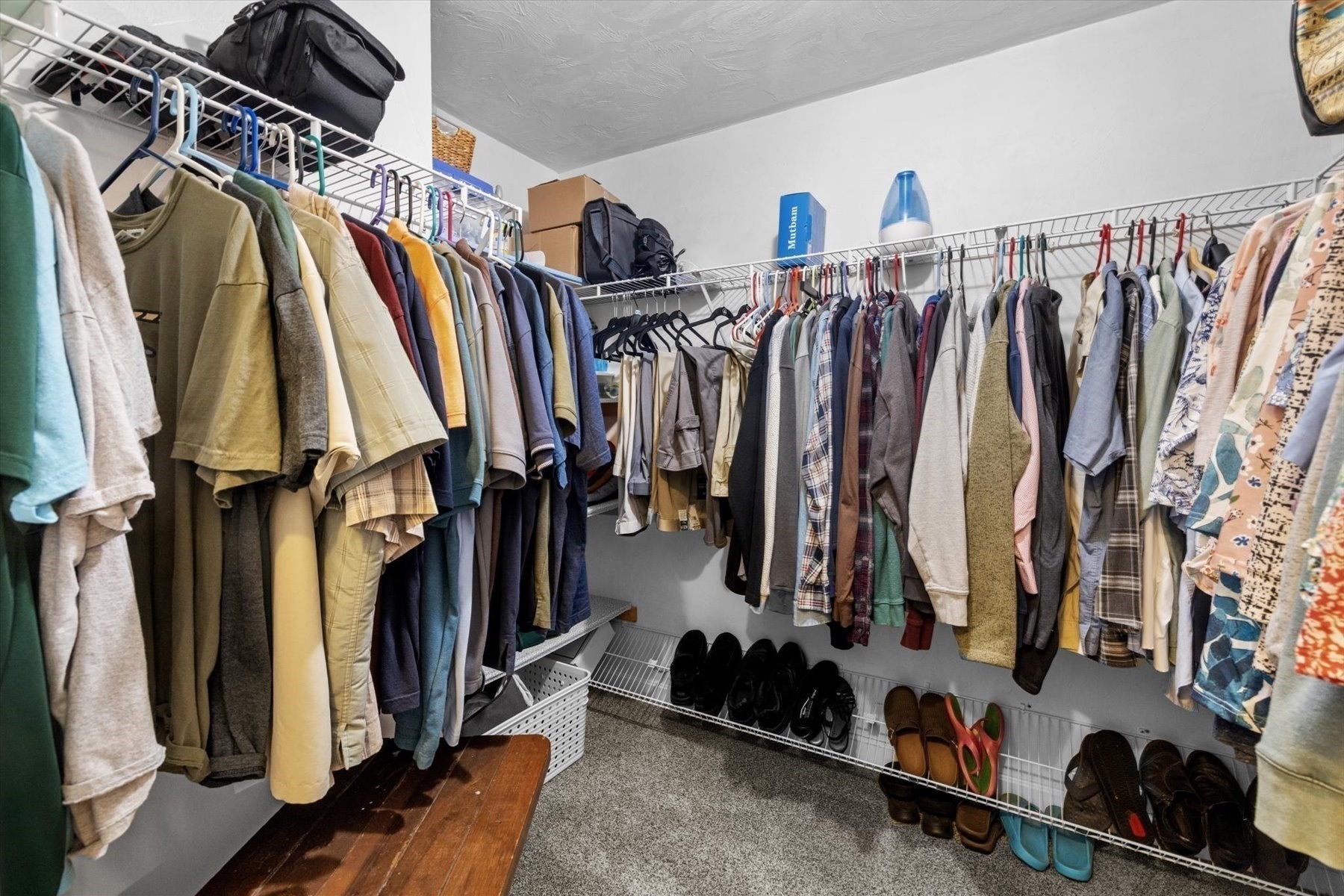 Walk-in closet with clothes on wire shelves, shoes below, and a wooden bench.
