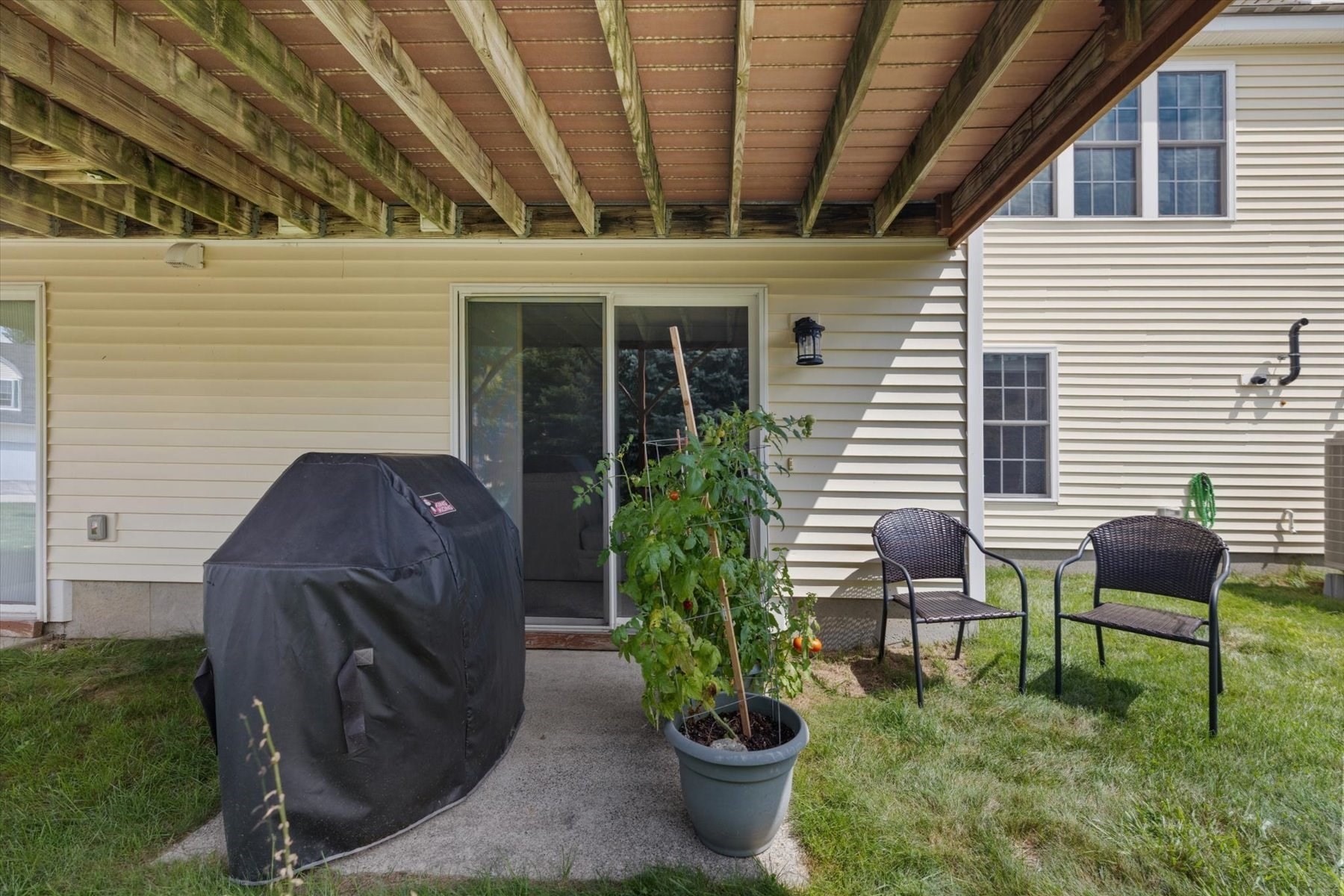Patio beneath a raised wooden deck with a covered grill, potted tomato plant, and two chairs.