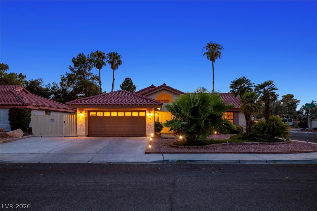 Single-story stucco home with red tile roof, illuminated entry, and palm landscaping at dusk.