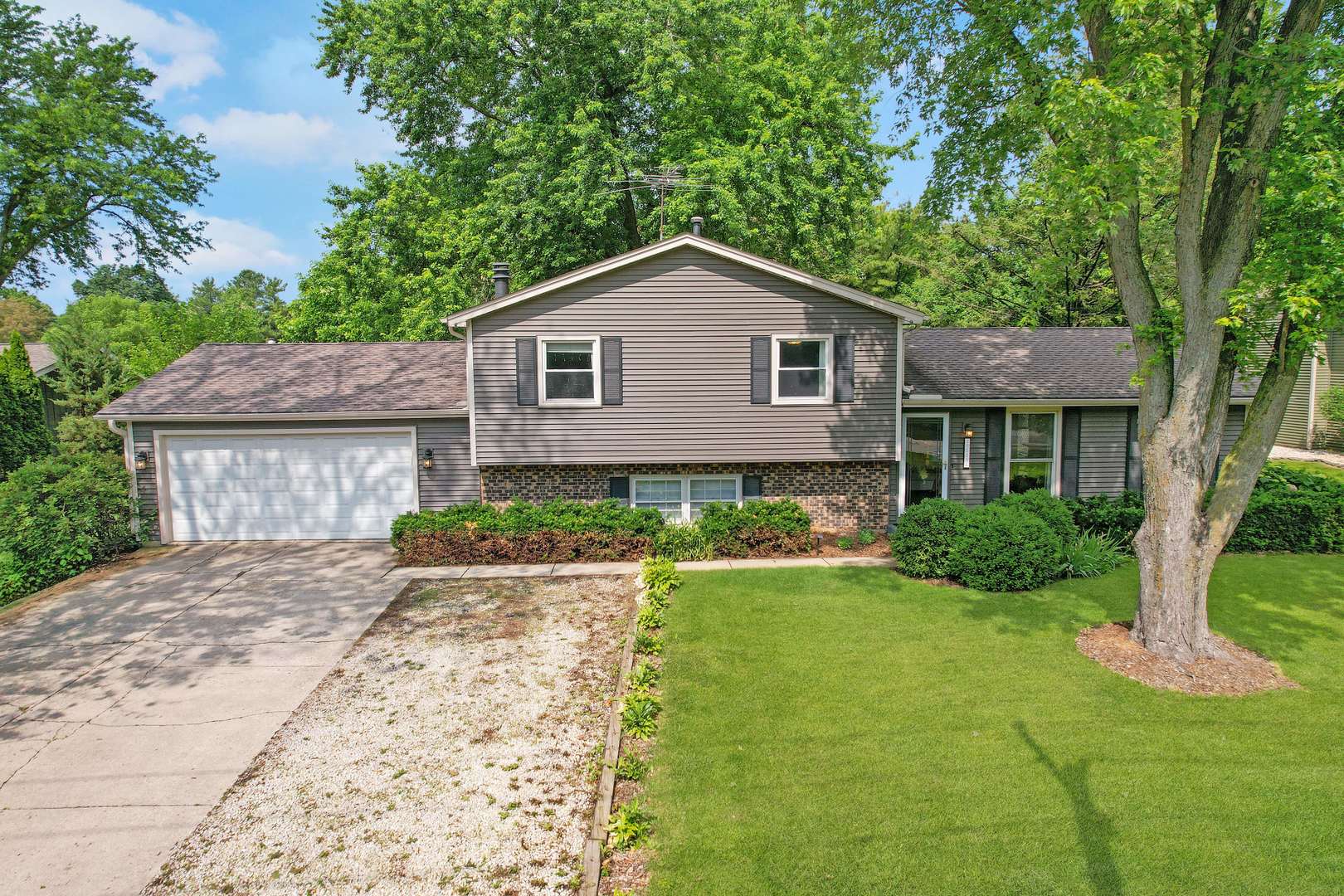 Gray split-level house with a two-car garage, brick lower facade, and mature trees.