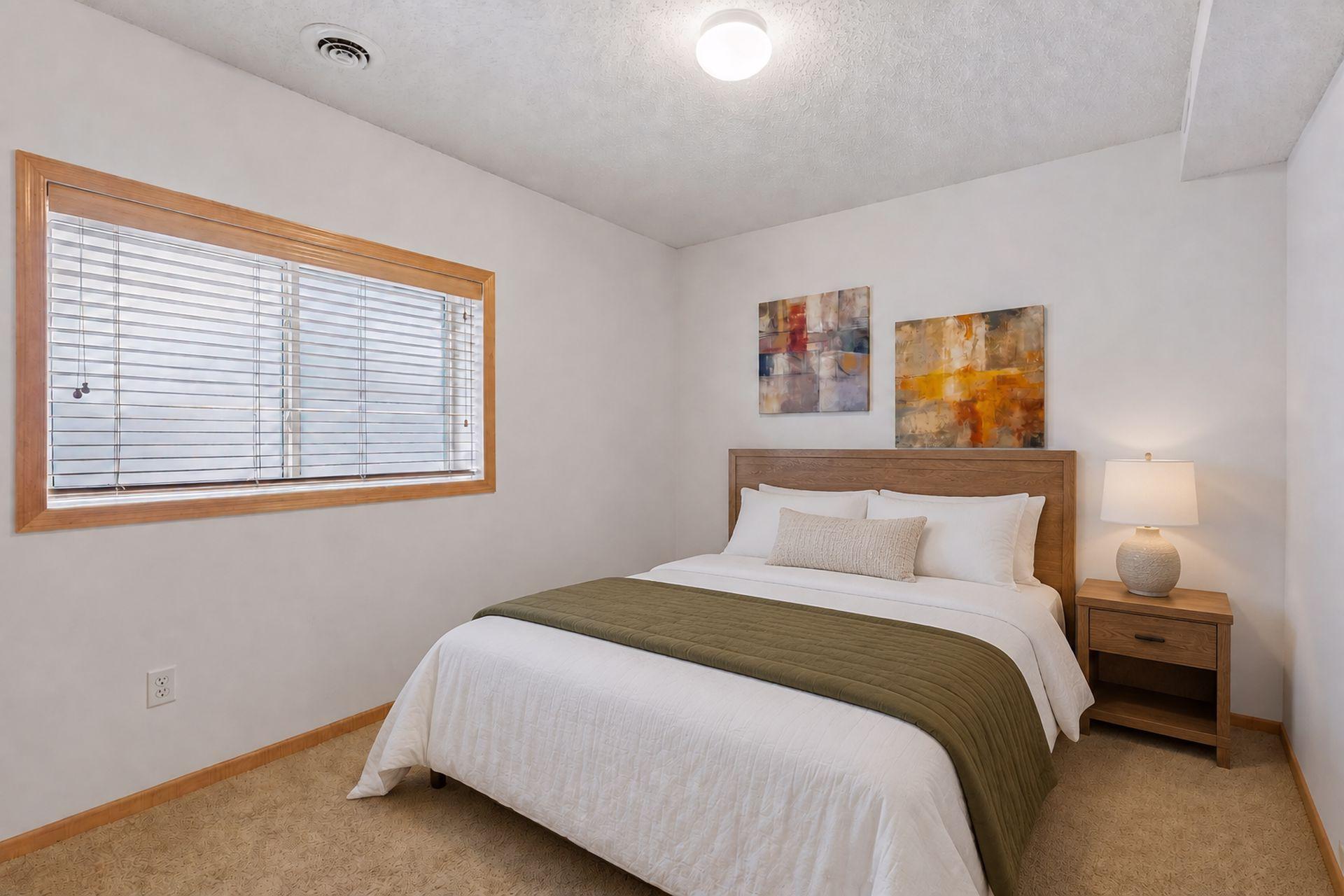 Staged bedroom with a white bed, olive-green quilt, wood furnishings, and a large window with blinds.