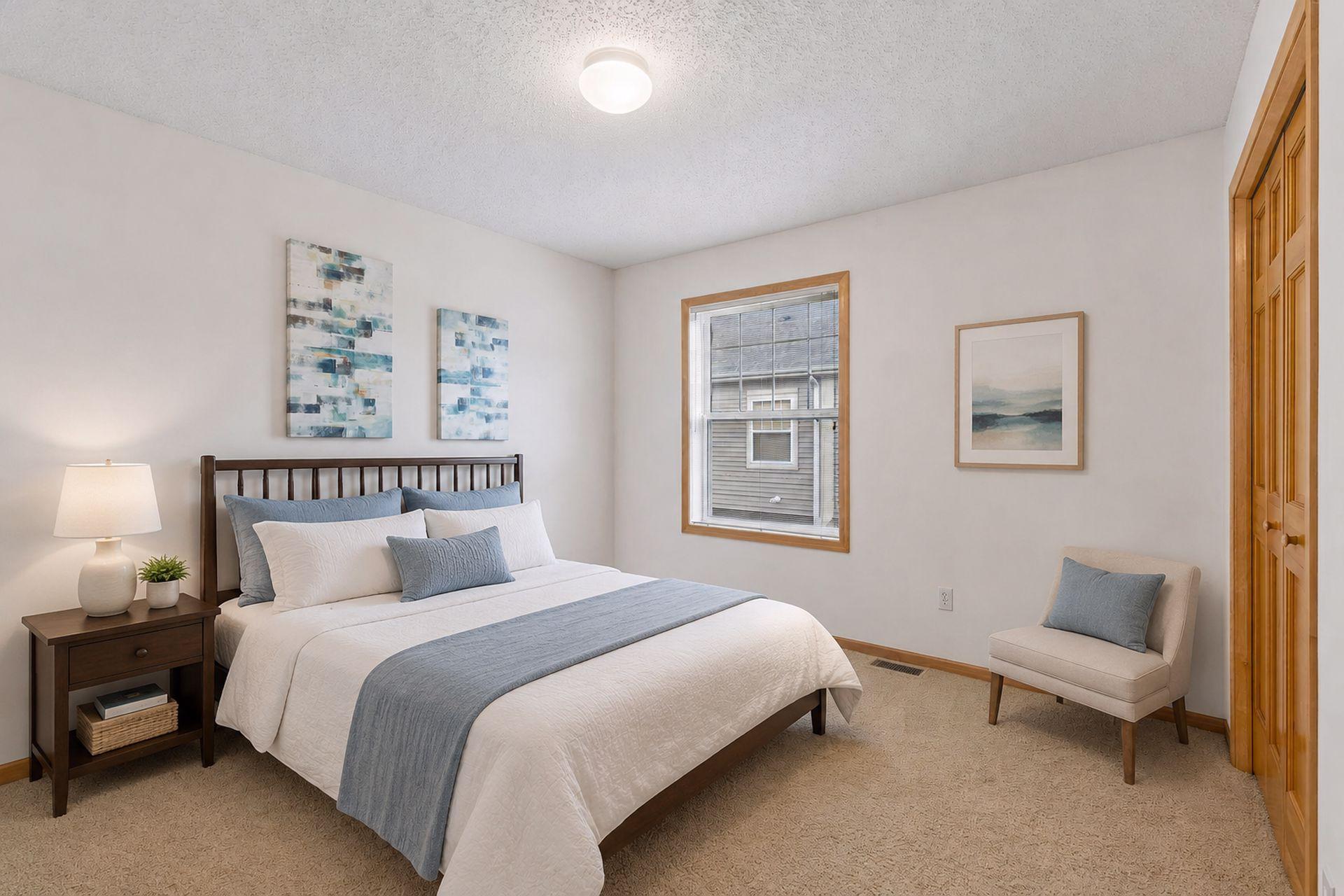 Staged bedroom with a white bed, blue-gray accents, wood-trimmed window, and cushioned chair.