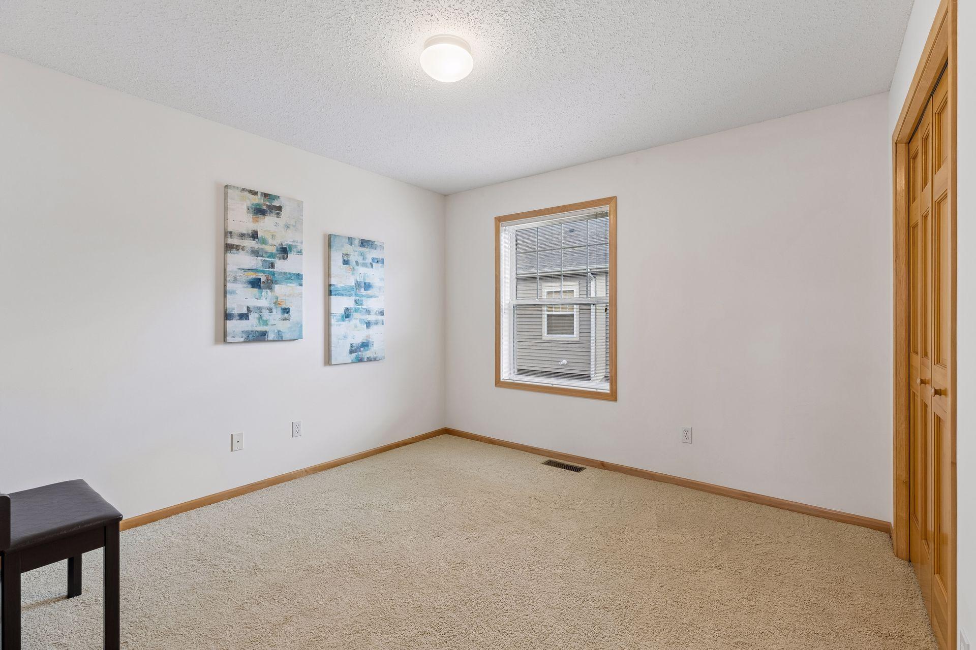 Mostly empty bedroom with beige carpet, a window, wood-trimmed closet doors, and abstract wall art.