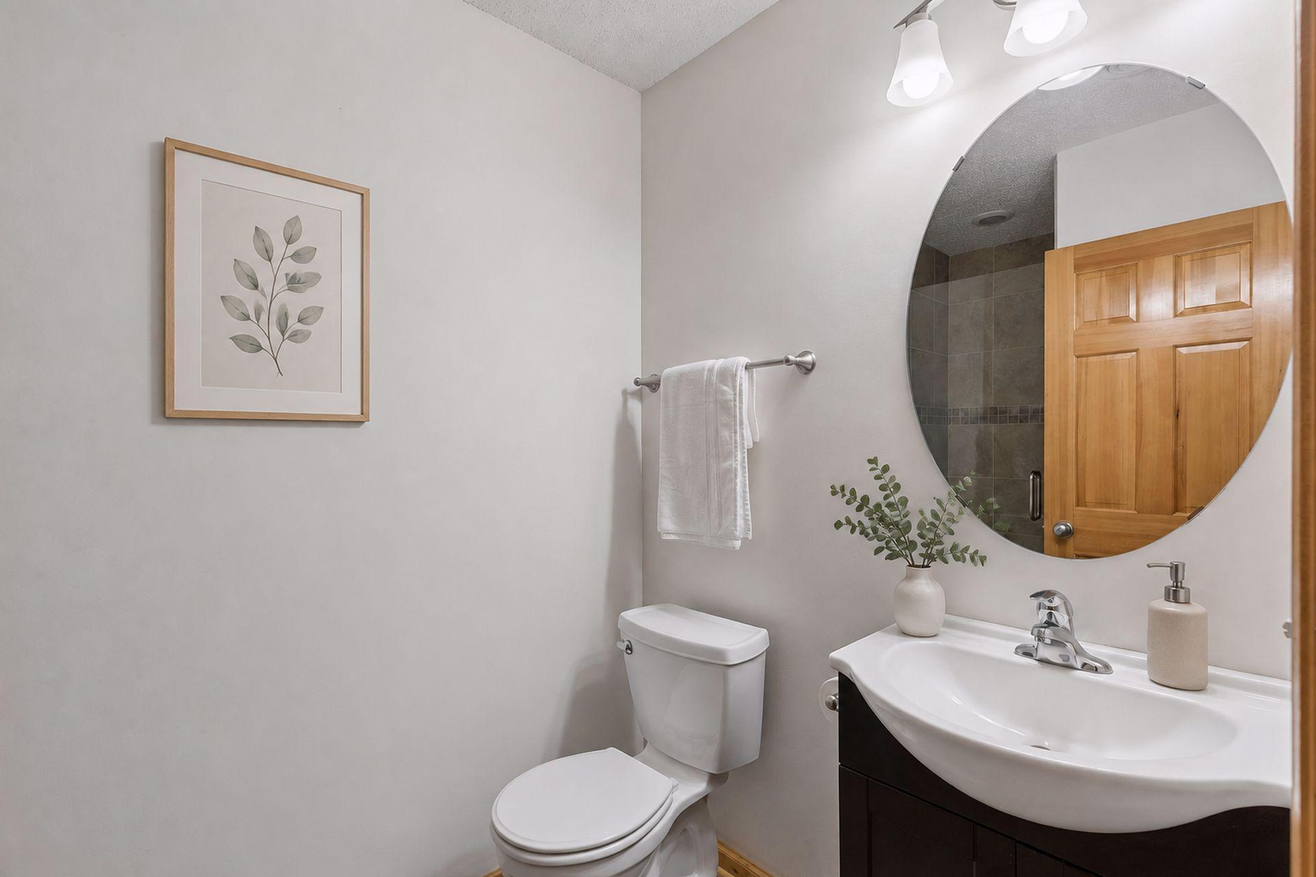 Bathroom with a round mirror, white sink and toilet, and a tiled shower reflected in the mirror.