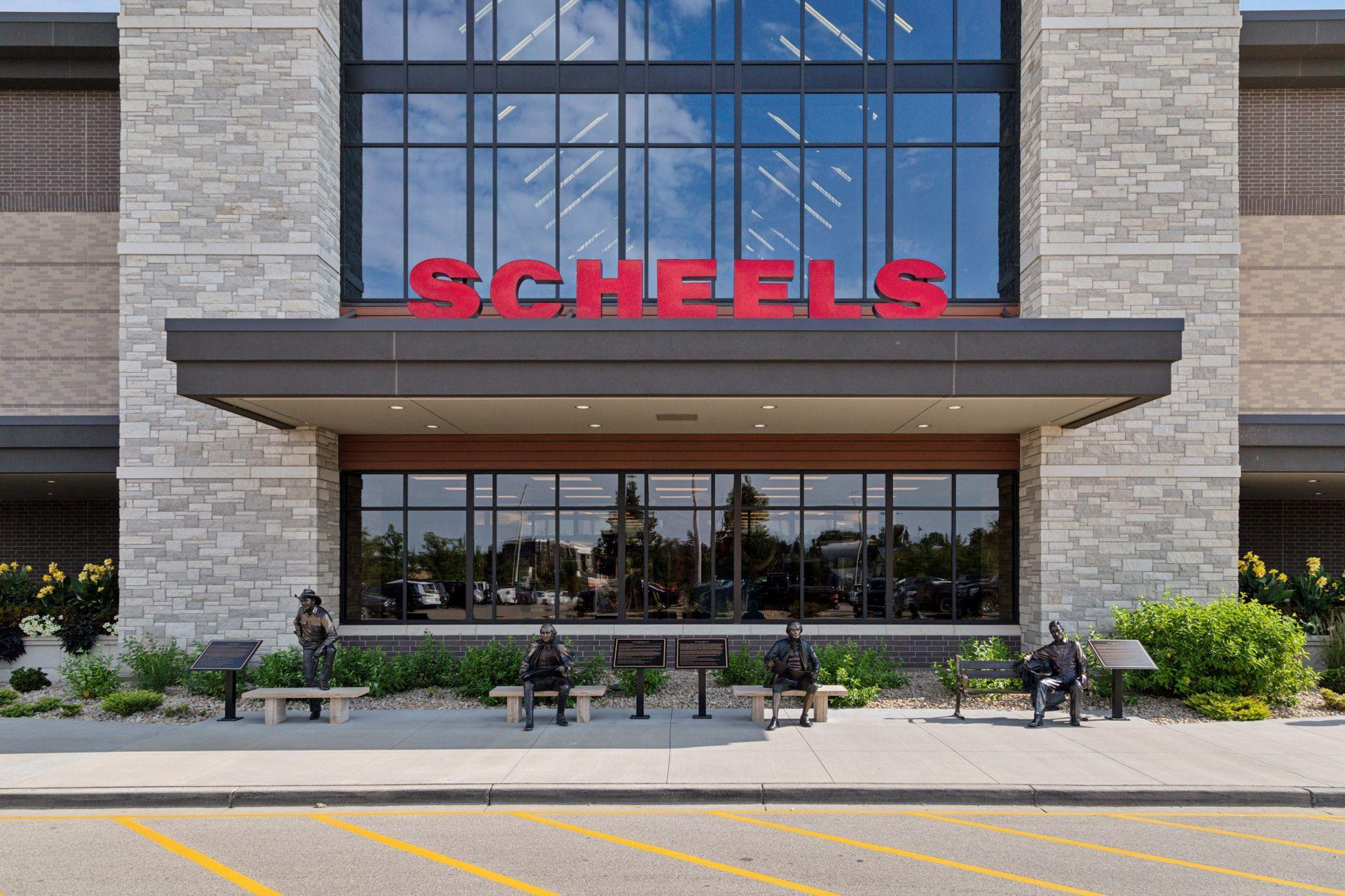 Scheels storefront with a large red sign and bronze statues seated outside.