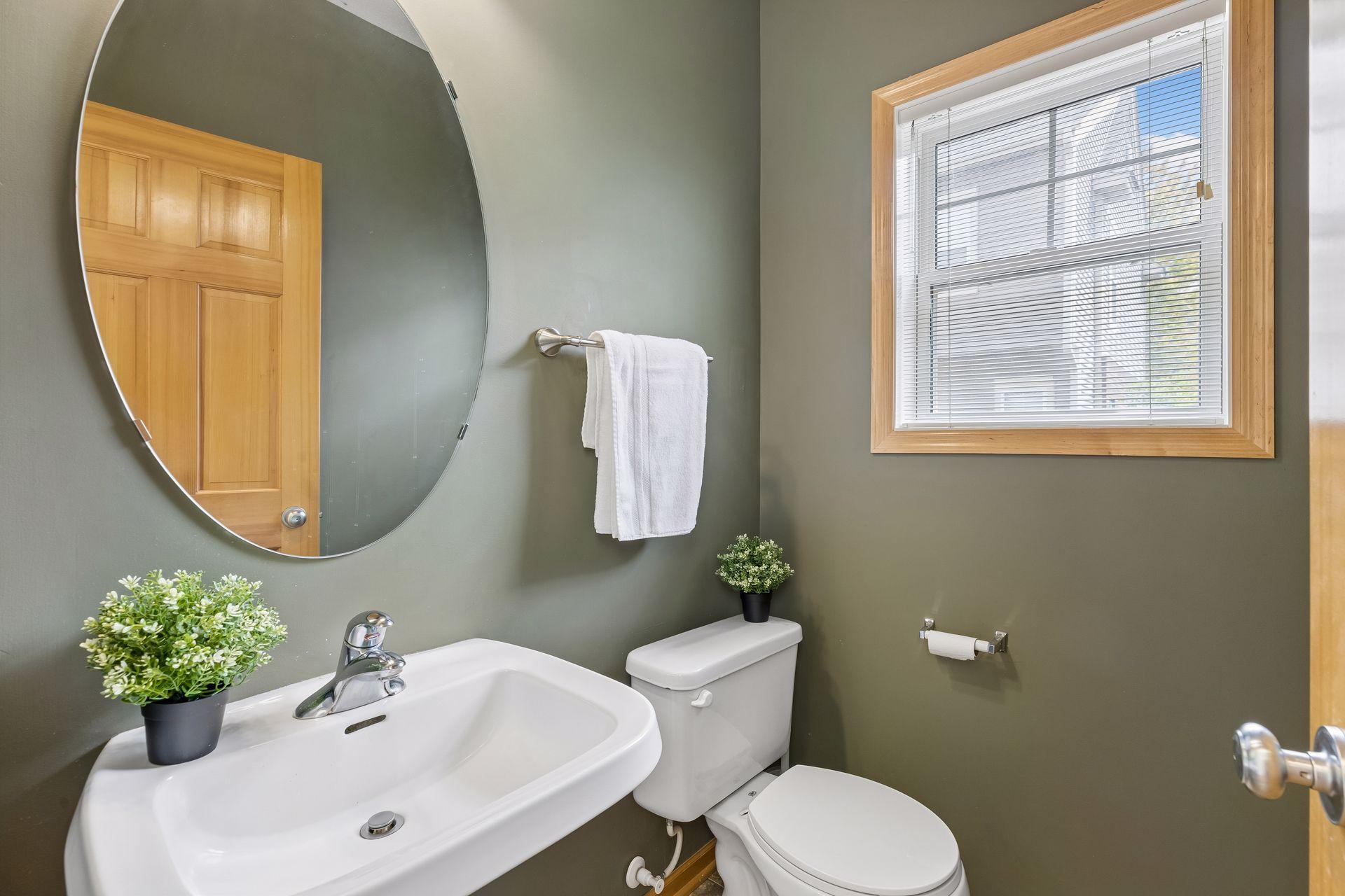 Bathroom with a large oval mirror, white sink and toilet, and a wood-trimmed window.