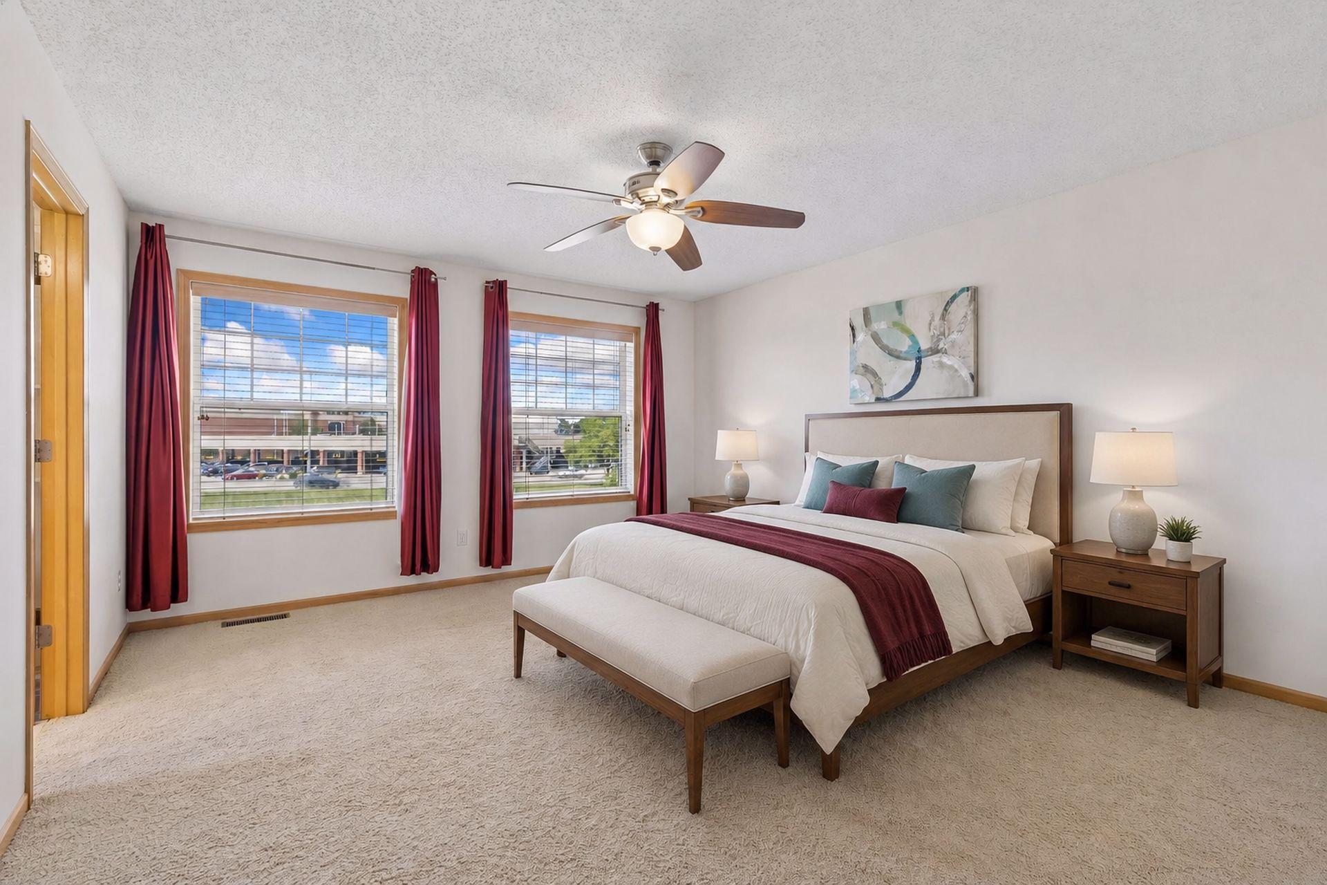 Furnished bedroom with a wood-framed bed, burgundy curtains, and two large windows.