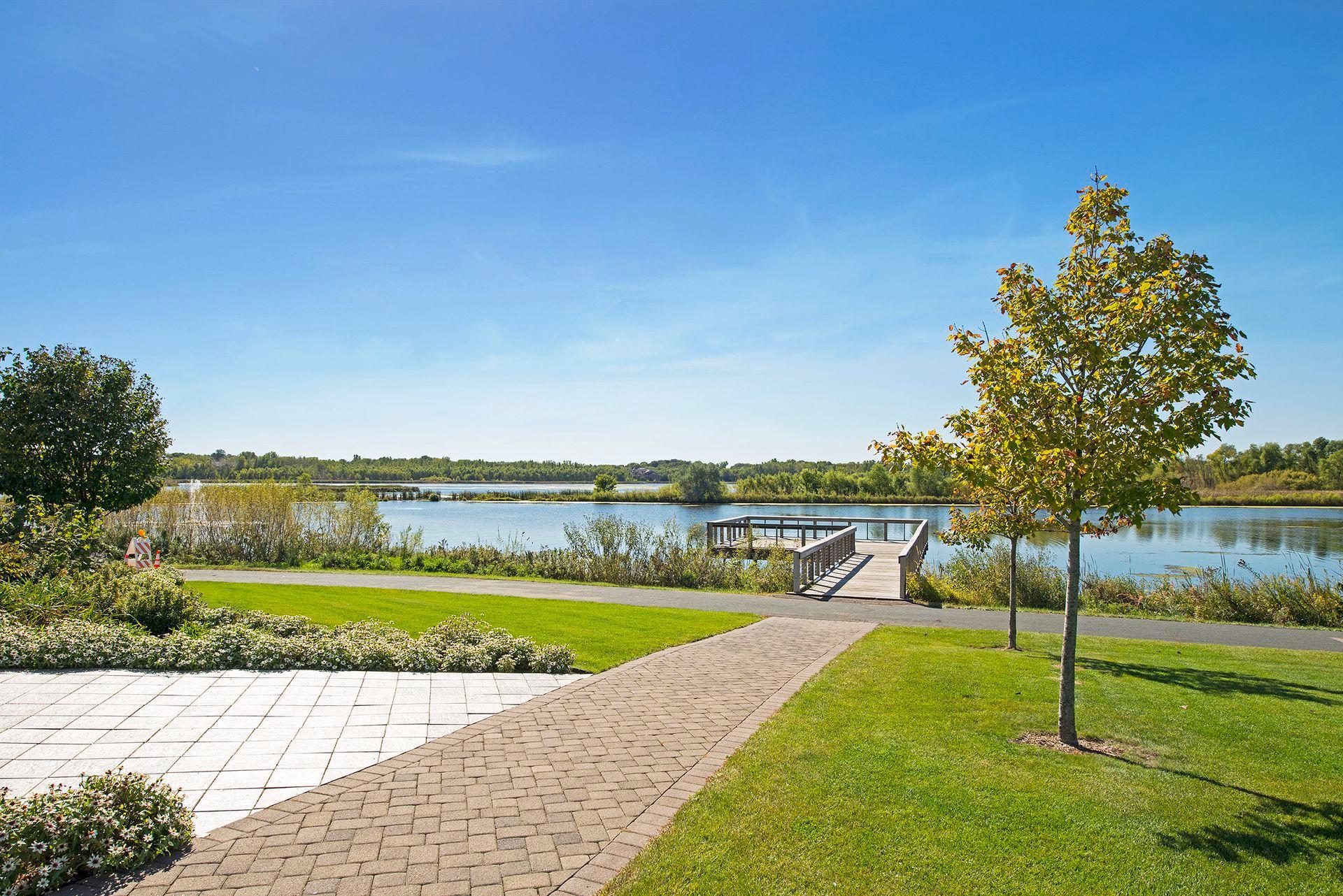 Landscaped waterfront with a wooden dock, paved path, and autumn-colored trees.