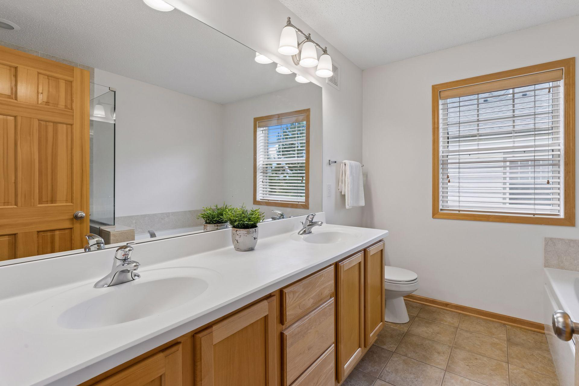Bathroom with a double-sink vanity, wood cabinetry, large mirror, and two windows with blinds.
