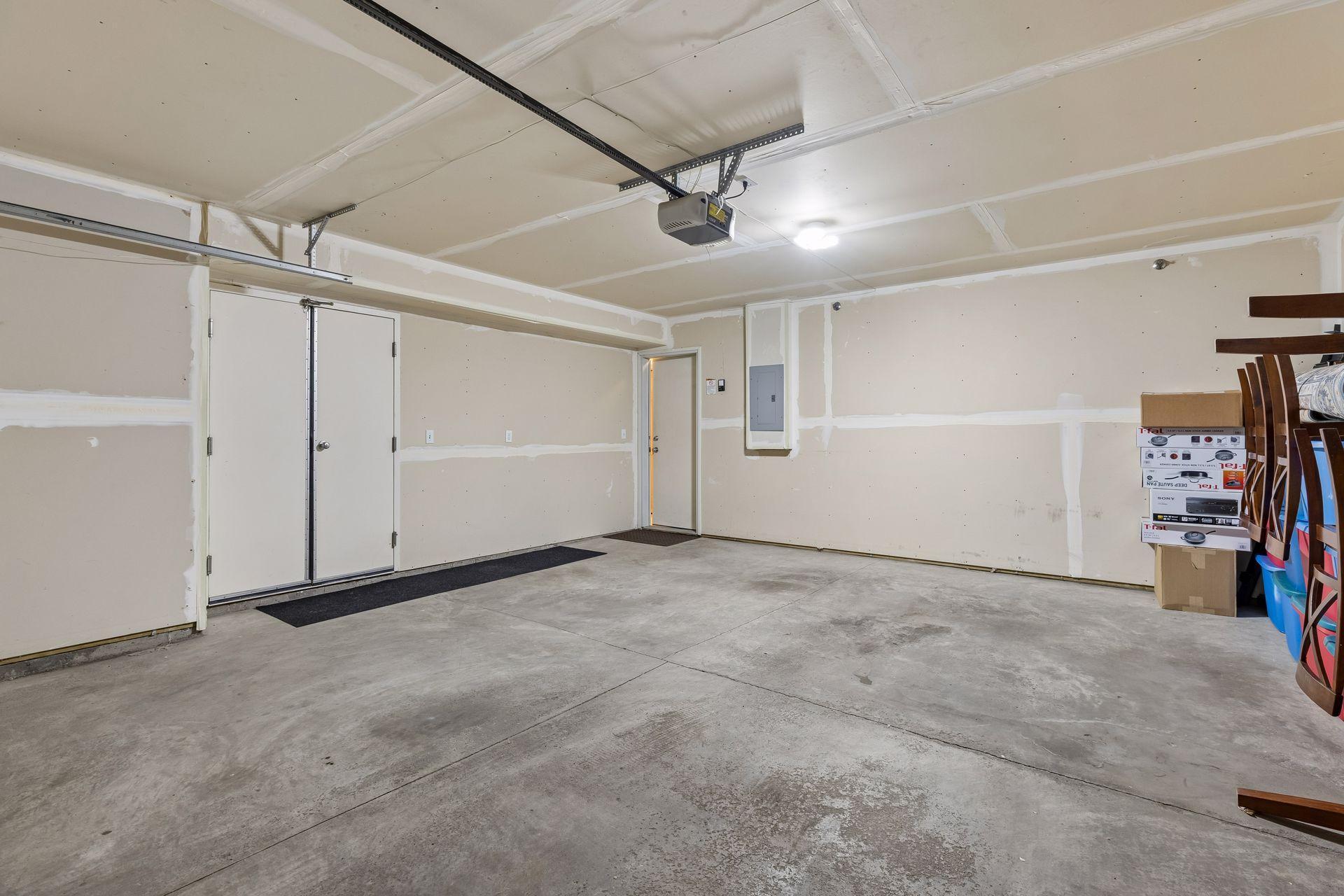 Garage with a concrete floor, unfinished drywall, and boxes and chairs stored along one wall.