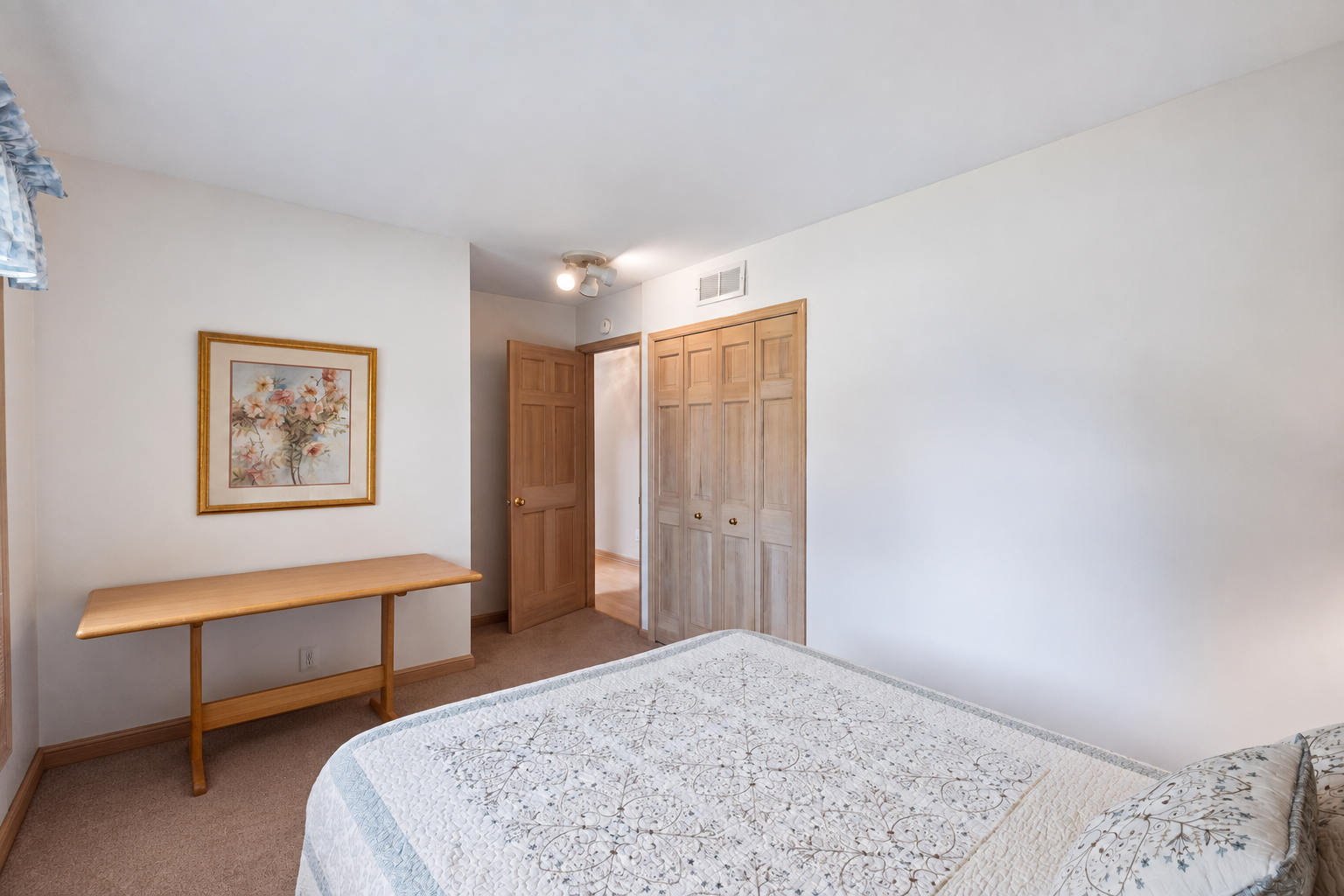 Bedroom with a patterned bedspread, wooden console table, floral artwork, and paneled closet doors.