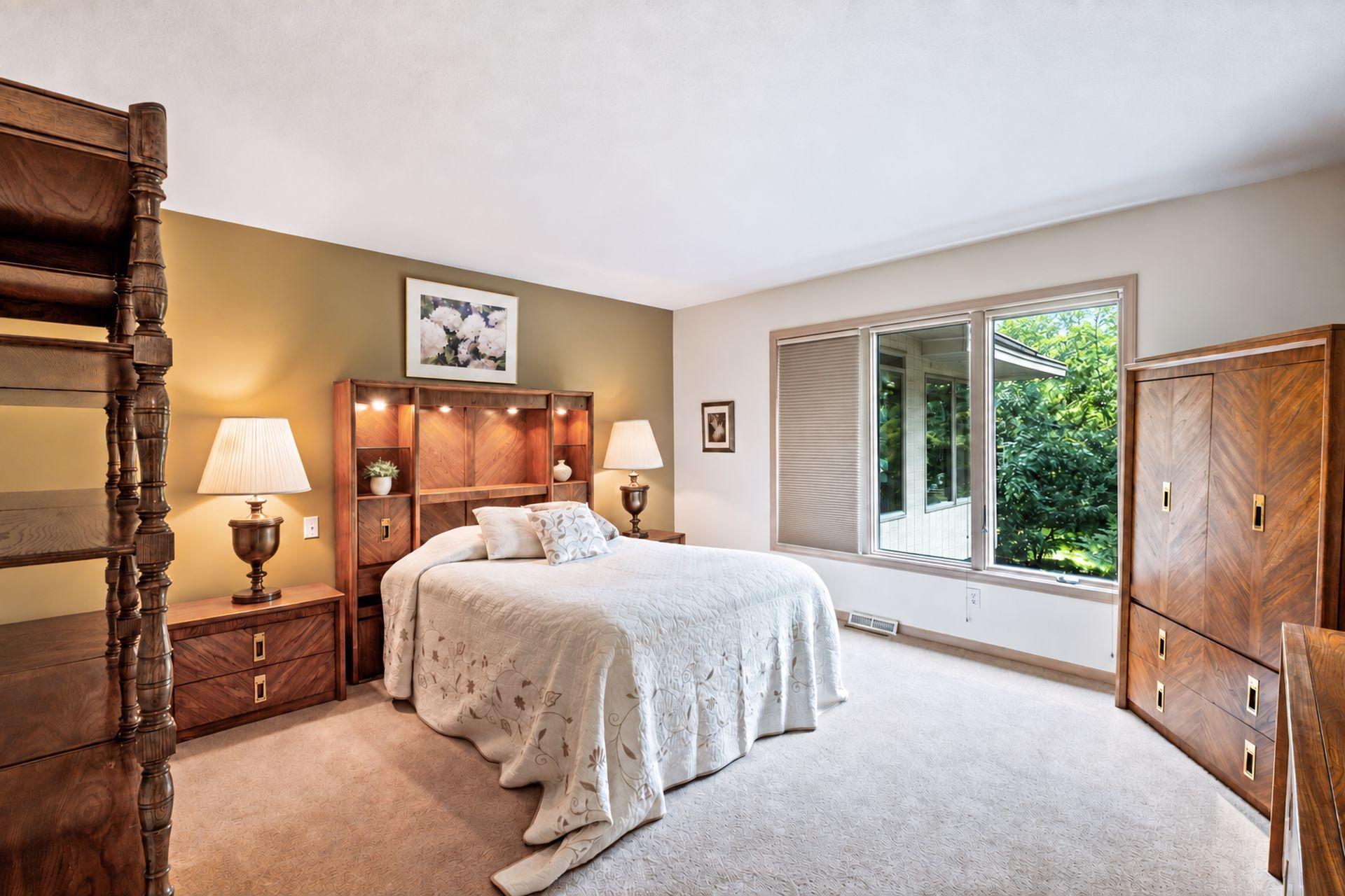 Bedroom with a large bed, matching dark wood furniture, and a wide window overlooking greenery.