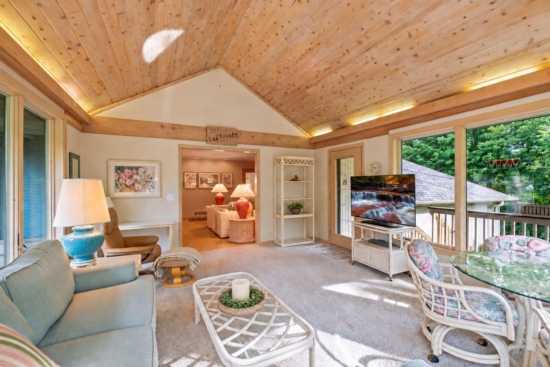 Sunroom with a vaulted wood ceiling, wicker seating, and large windows opening toward a tree-lined deck.