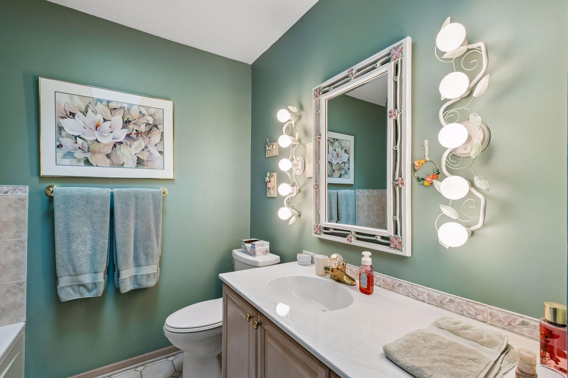 Bathroom with sage-green walls, a white vanity, floral-framed mirror, and ornate wall lights.