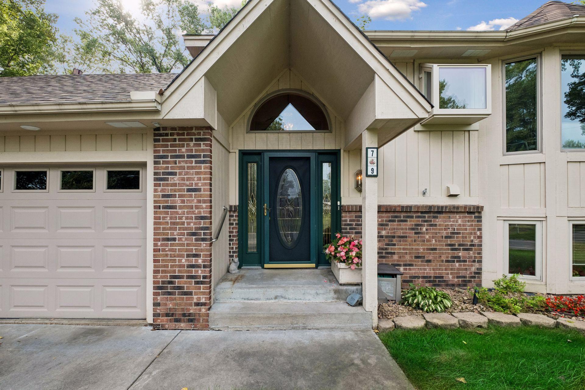 House entrance with a dark green oval-window door, peaked covered entry, brick accents, and attached garage.