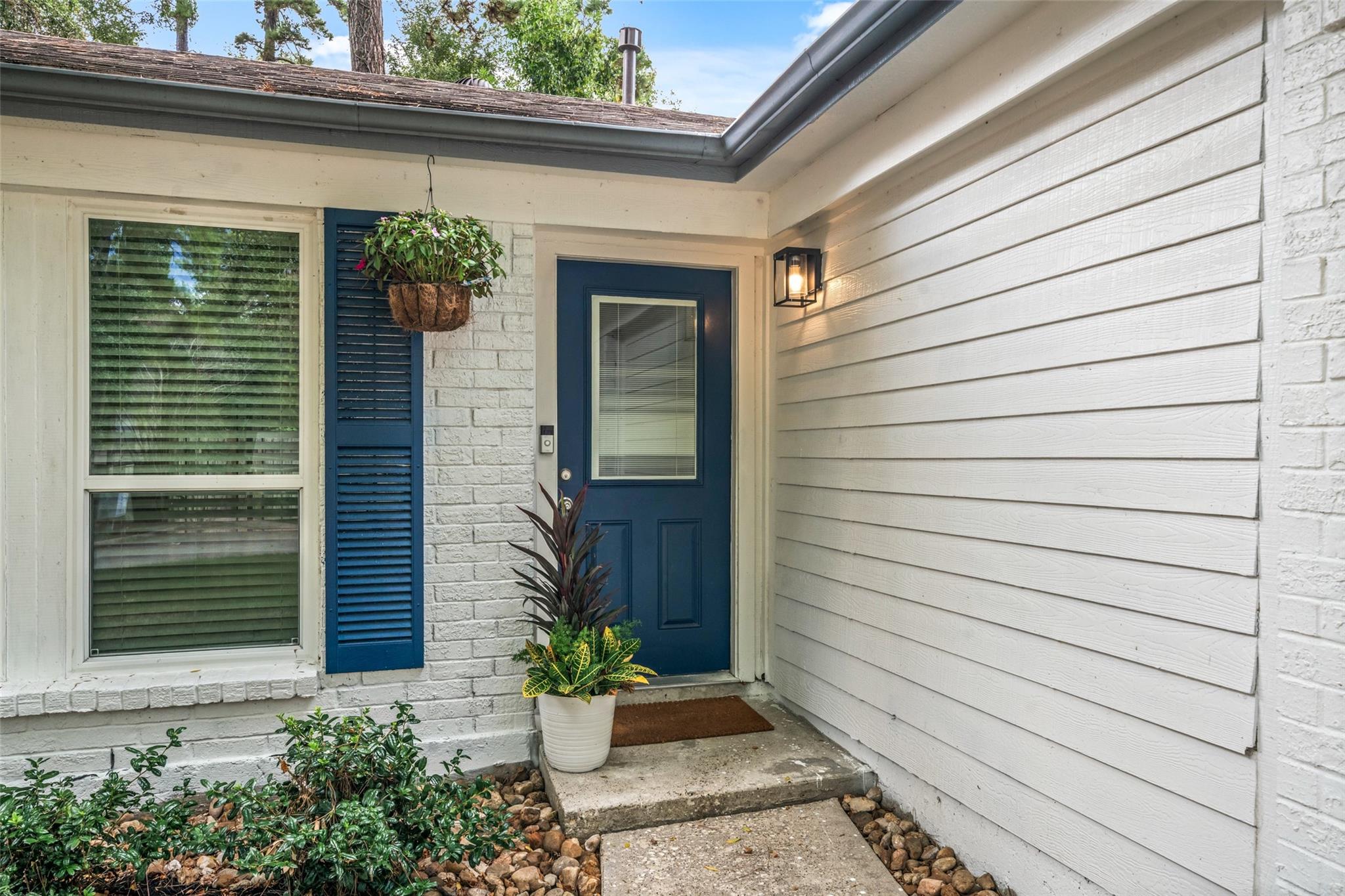 Covered home entrance with a dark blue door, white siding, blue shutters, and potted plants.