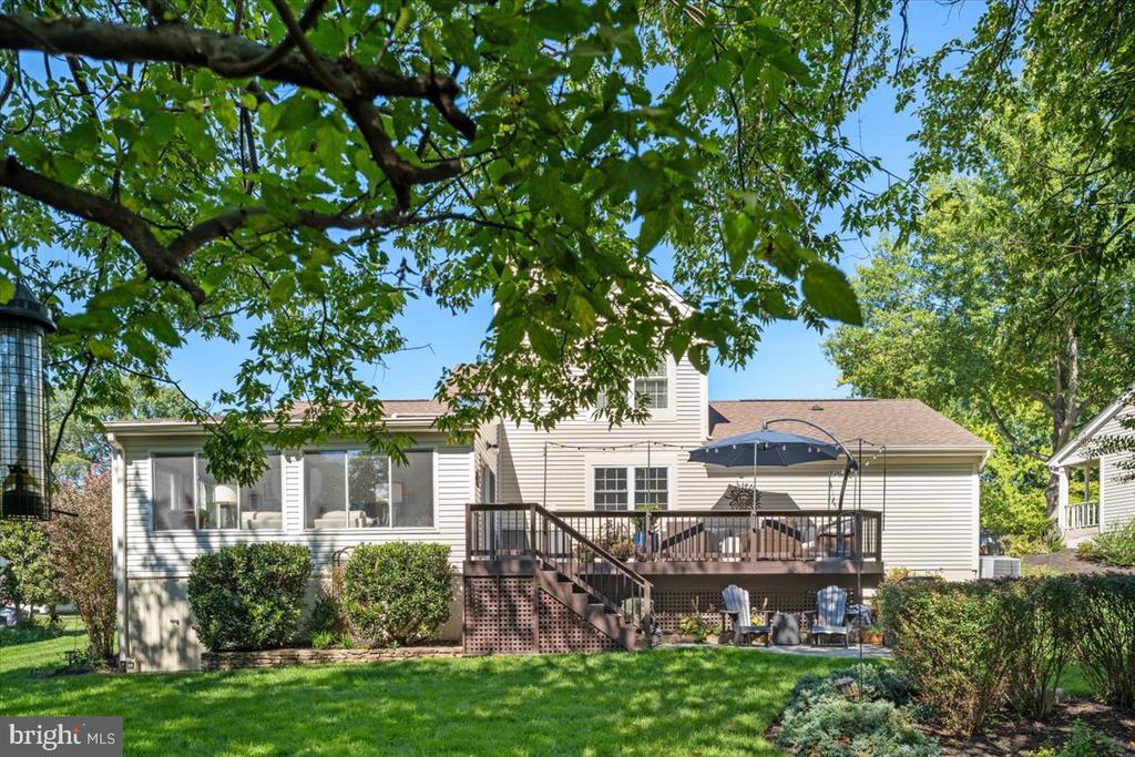Rear of a light-colored home with a raised deck and umbrella beneath mature trees.