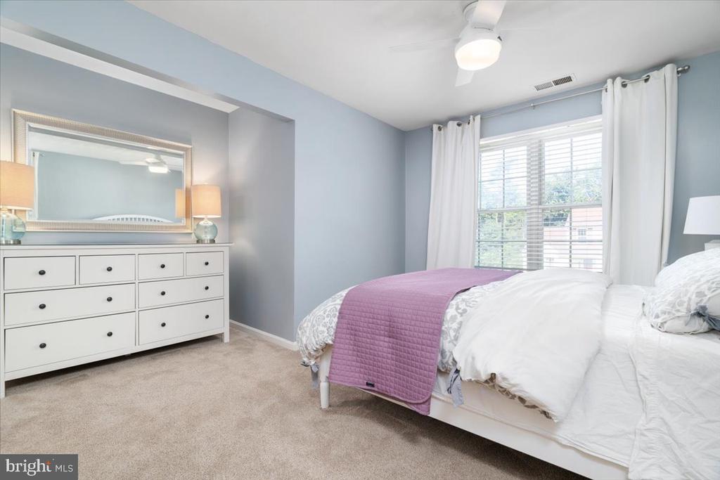 Furnished bedroom with a white bed, mauve quilt, mirrored dresser, and bright window.