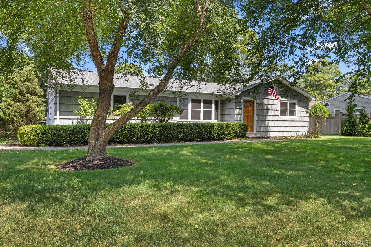 Gray shingle-sided single-story house with a wooden front door, American flag, and mature tree in the front yard.