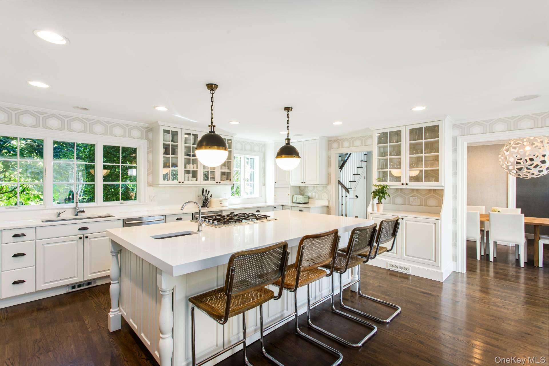 Bright white kitchen with a large island, woven-seat stools, glass-front cabinets, and windows overlooking greenery.