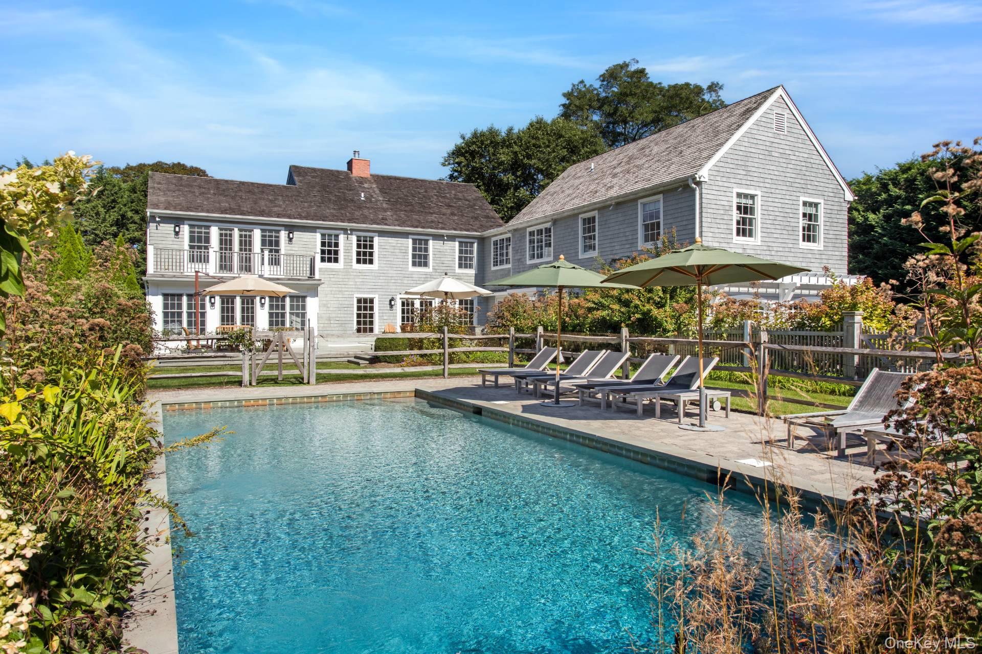 Backyard swimming pool beside a gray shingle home, with patio loungers and umbrellas.